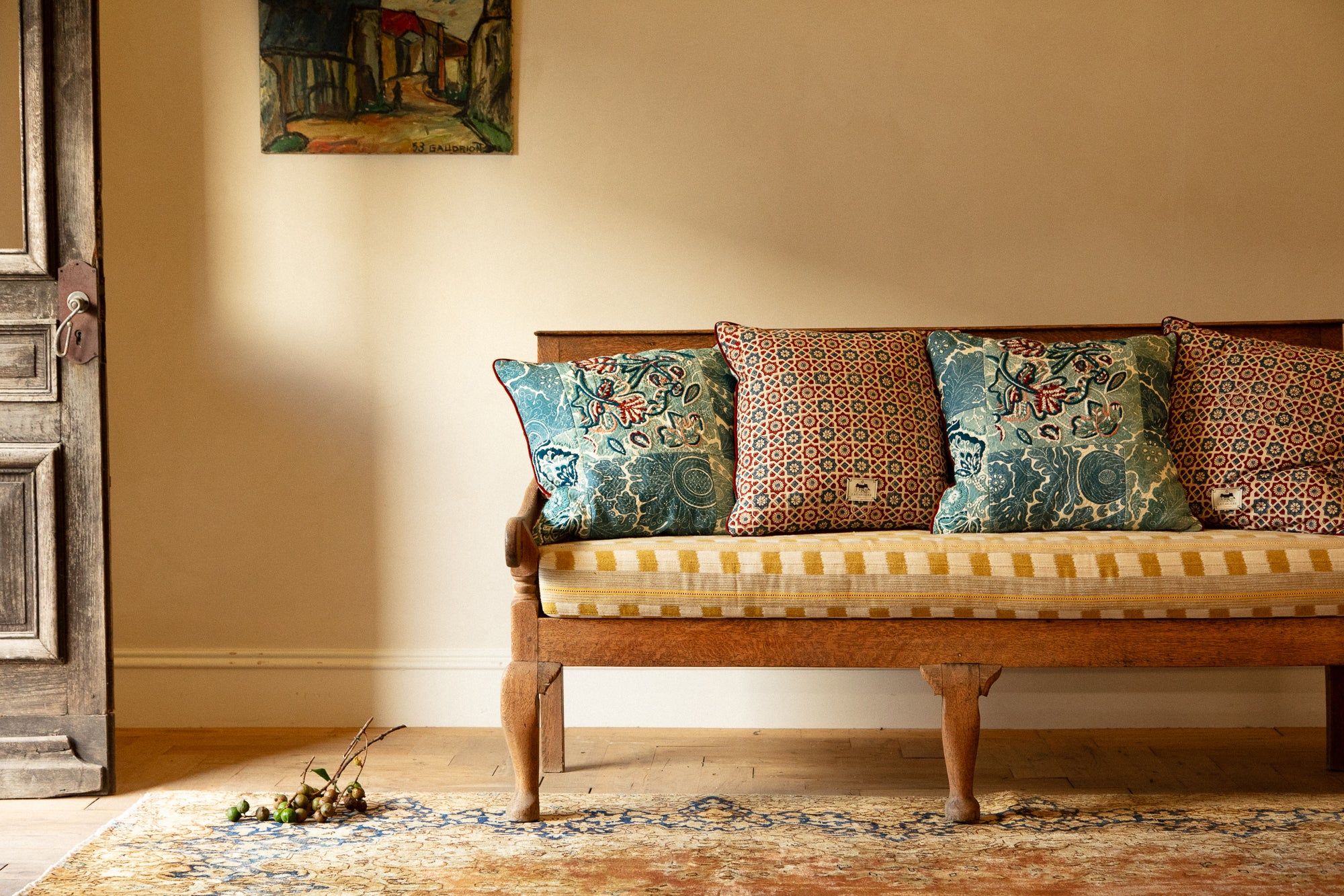 Wooden bench with striped cushion, patterned pillows. Branch near open rustic door. Colourful painting on cream wall above bench.