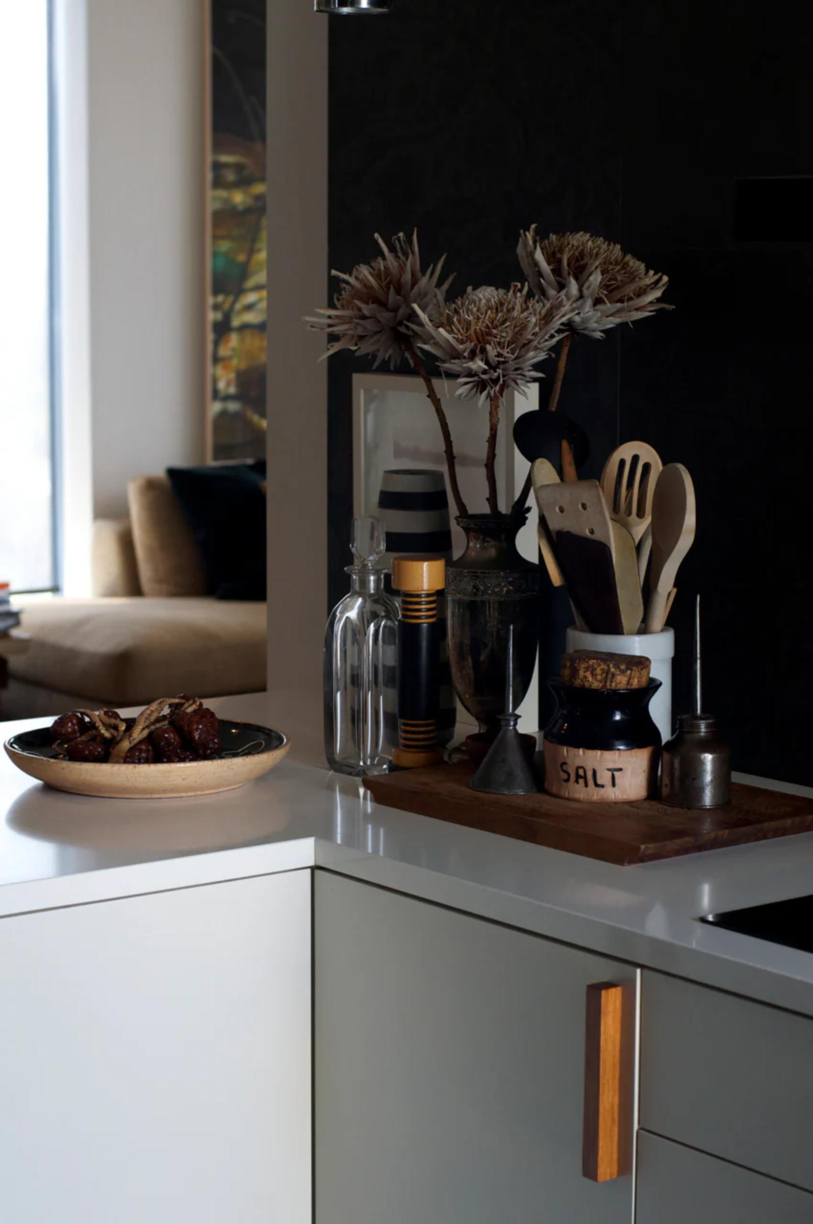 Minimalist kitchen corner with fruit bowl, utensils, and decorative items on counter. Beige sofa visible near large window in background.