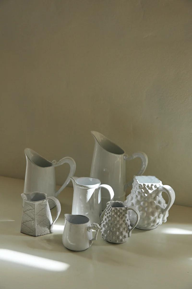 Various white ceramic pitchers on light surface. Different sizes and designs. Soft shadows, neutral wall background.