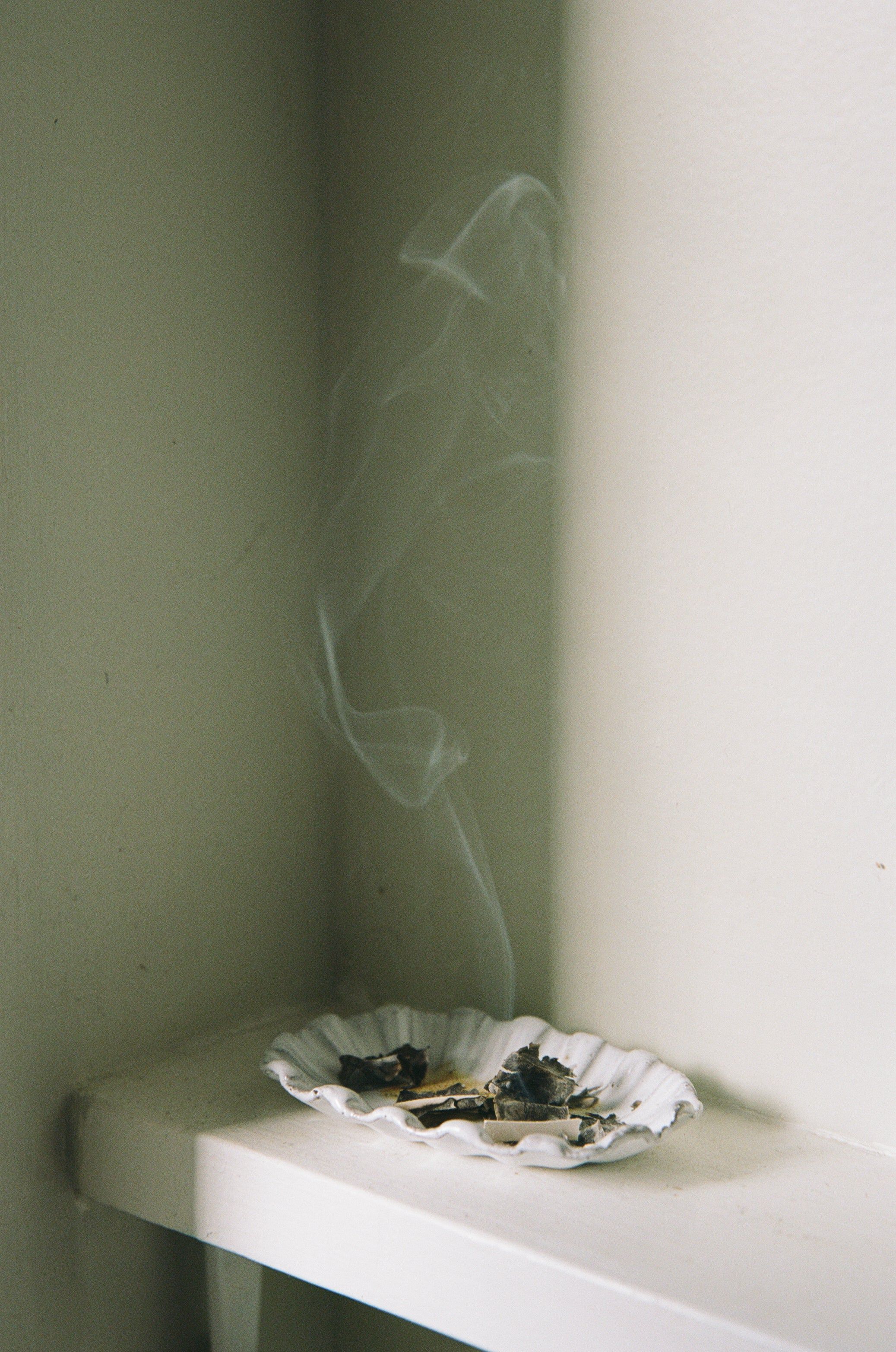 A white ceramic plate on a shelf holds burnt paper and ashes, with smoke rising gracefully against a light-coloured wall from burning benzoin.