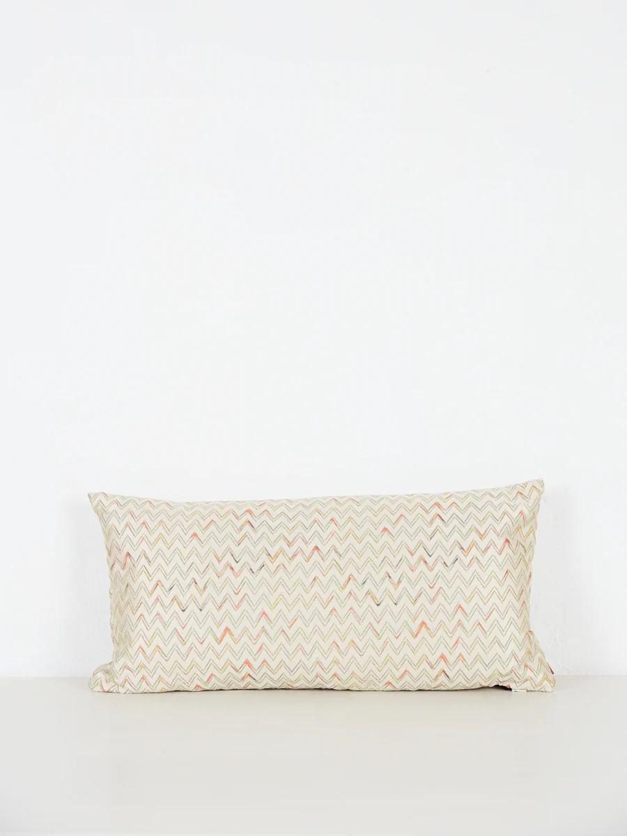 Rectangular chevron pillow in beige, orange, light brown. Against white wall on light surface.