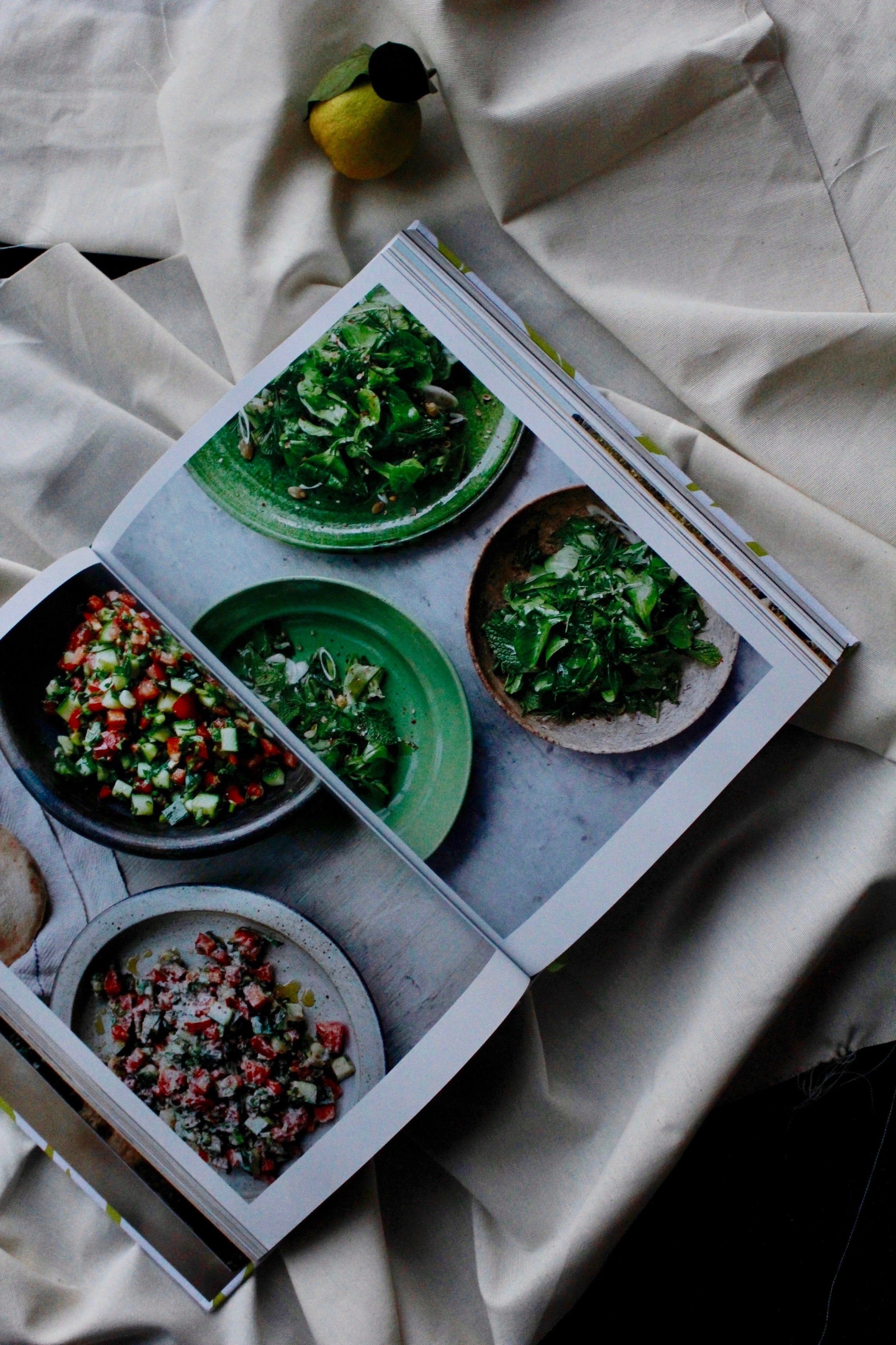 An open cookbook shows colourful salad dishes in green and brown bowls on a rippled light cloth. A yellow gourd lies nearby, evoking the _Falastin_ cookbook style.