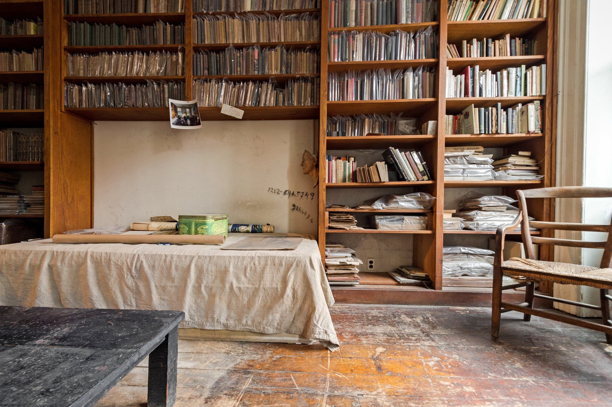 Library with tall bookshelves and pen-portraits. Central table with various items on beige cloth. Wooden chair and worn floor add rustic charm.