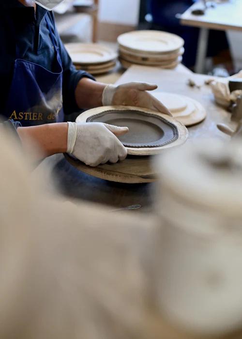 Person wearing gloves forming ceramic plate on pottery wheel. Finished and unfinished plates in background. Face partially obscured by mask.