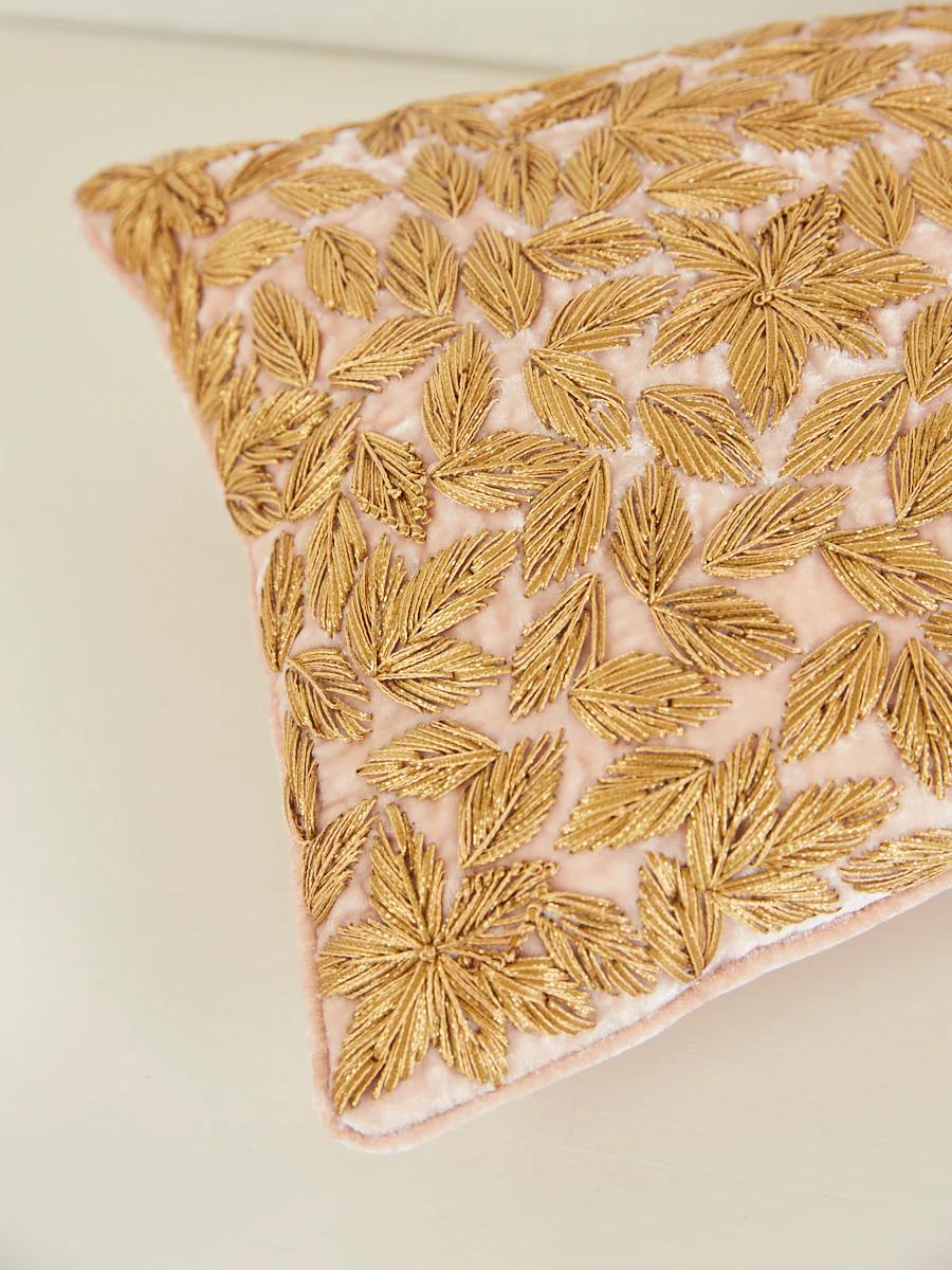 A decorative pillow with a light pink background is adorned with intricate gold leaf embroidery patterns. The close-up image highlights the detailed texture and craftsmanship.