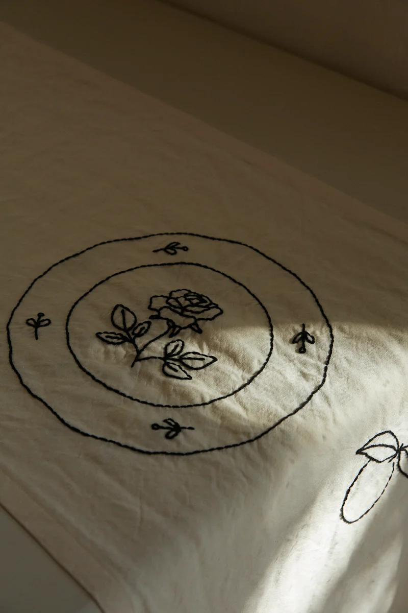 Off-white fabric with embroidered rose surrounded by circles and botanical elements. Soft shadows visible.