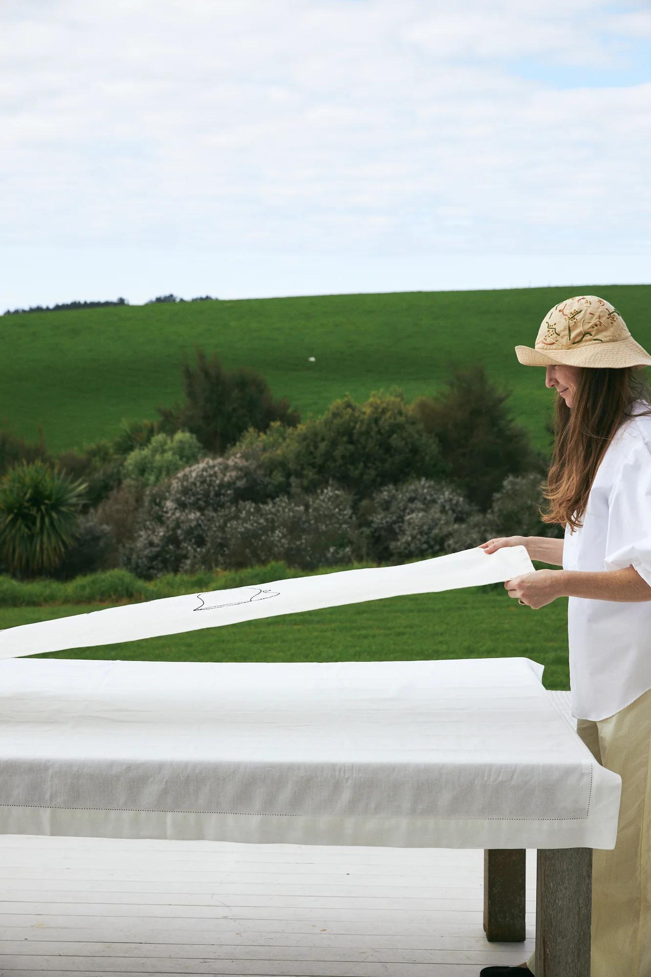 Woman in hat setting white tablecloth on outdoor table. Green fields and trees under partly cloudy sky.