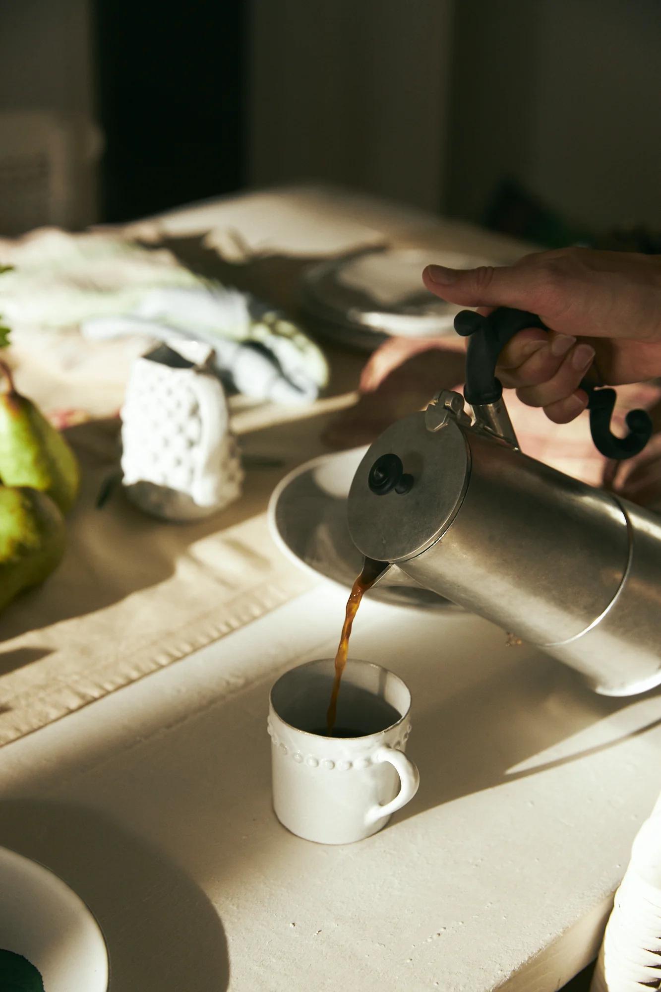 Person pouring coffee from stovetop maker into textured mug. Light-coloured table with pears and ceramic owl in background.