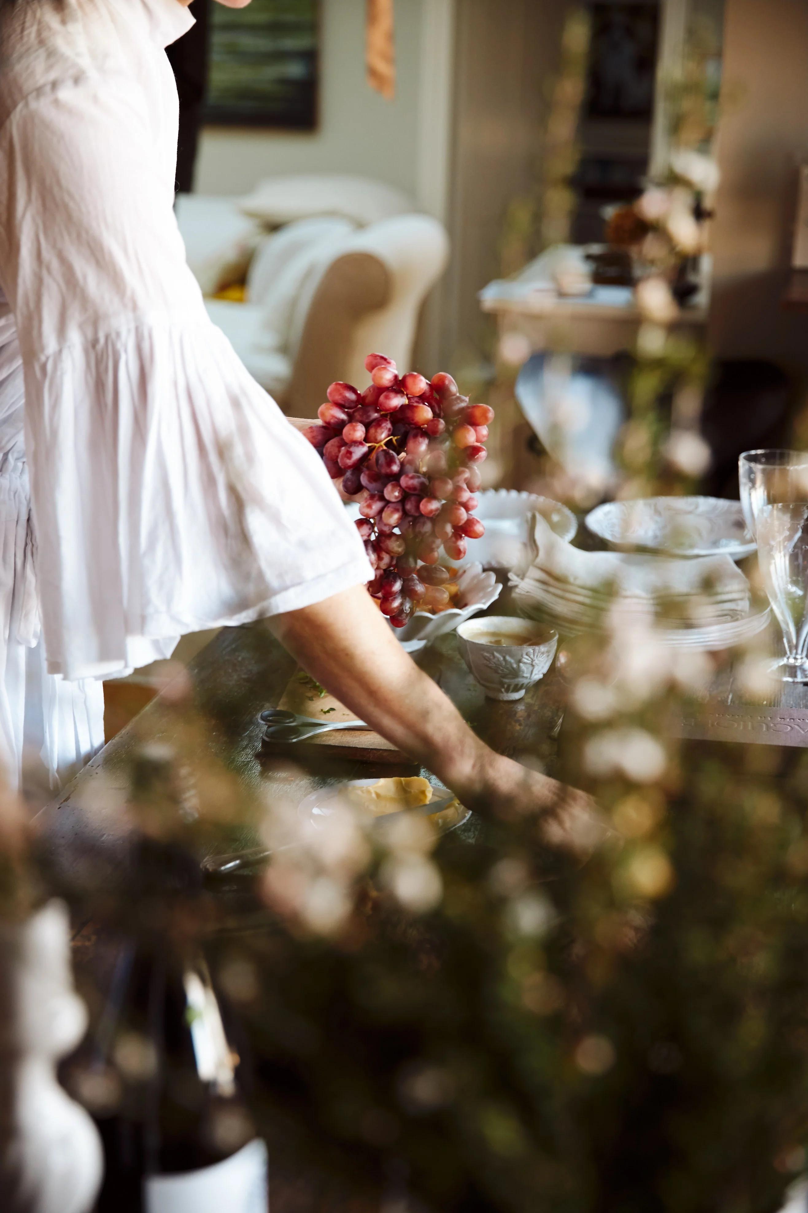 Person in white shirt setting table with grapes. Greenery in foreground, cream sofa in background.