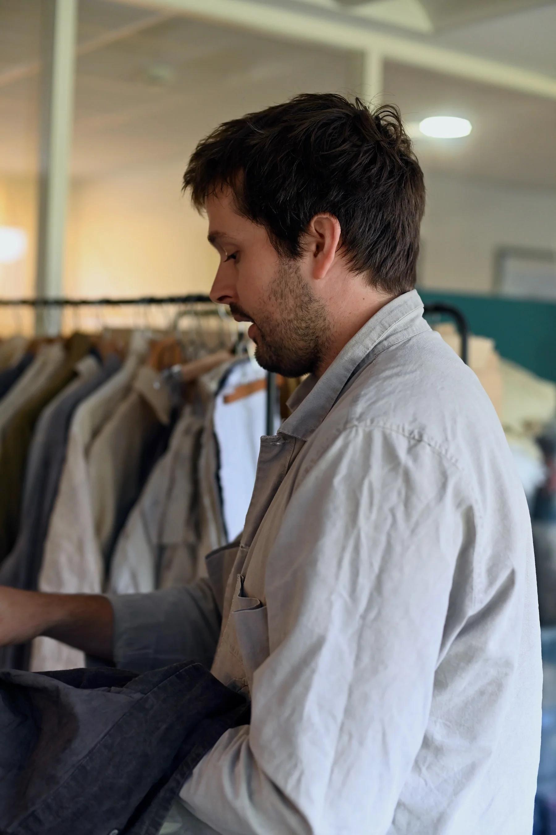 Man with dark hair examining clothes on rack. Light-coloured shirt, indoor setting.