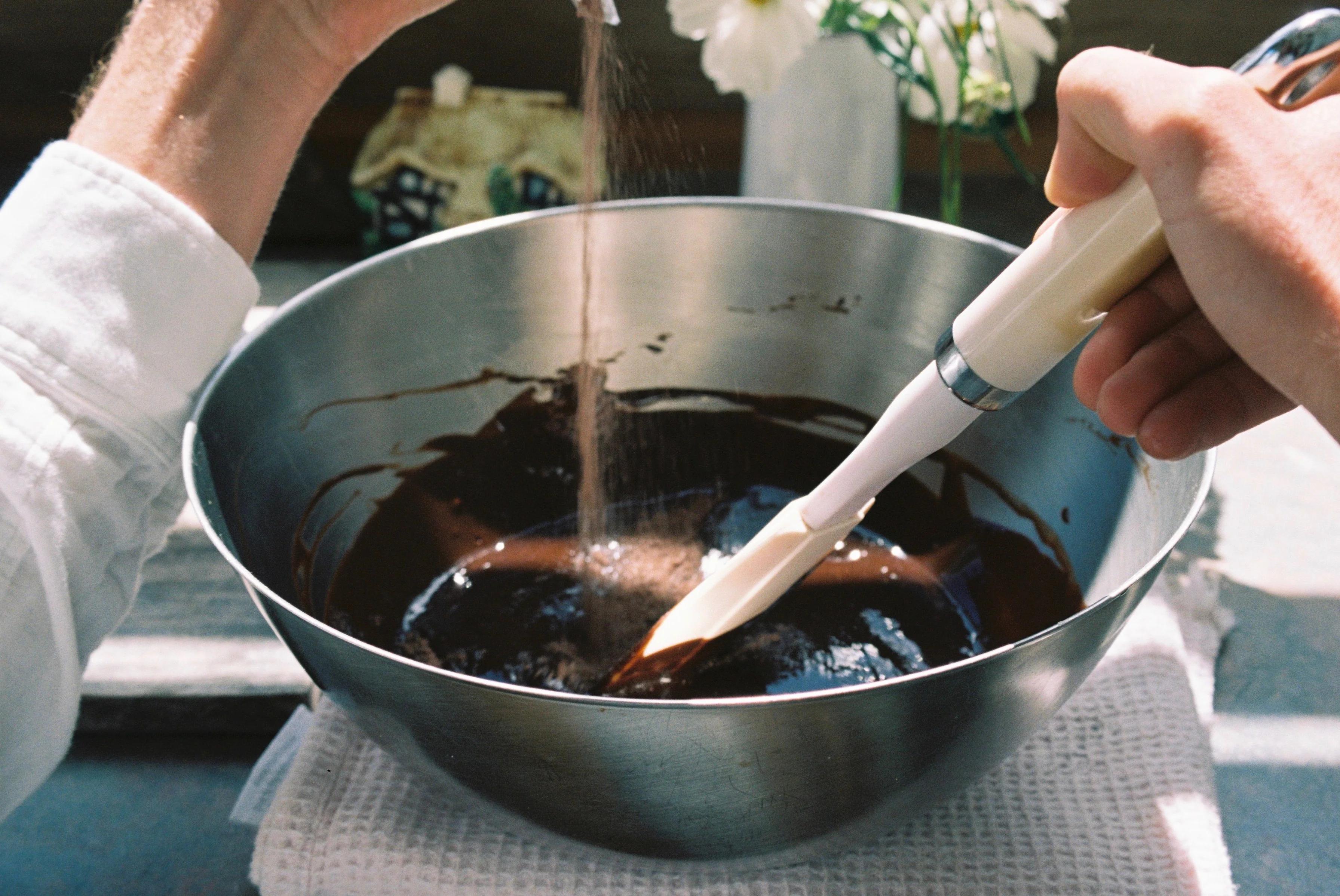 A person sprinkling sugar into chocolate batter in a large bowl while another hand mixes with a white spatula, preparing a festive cake.