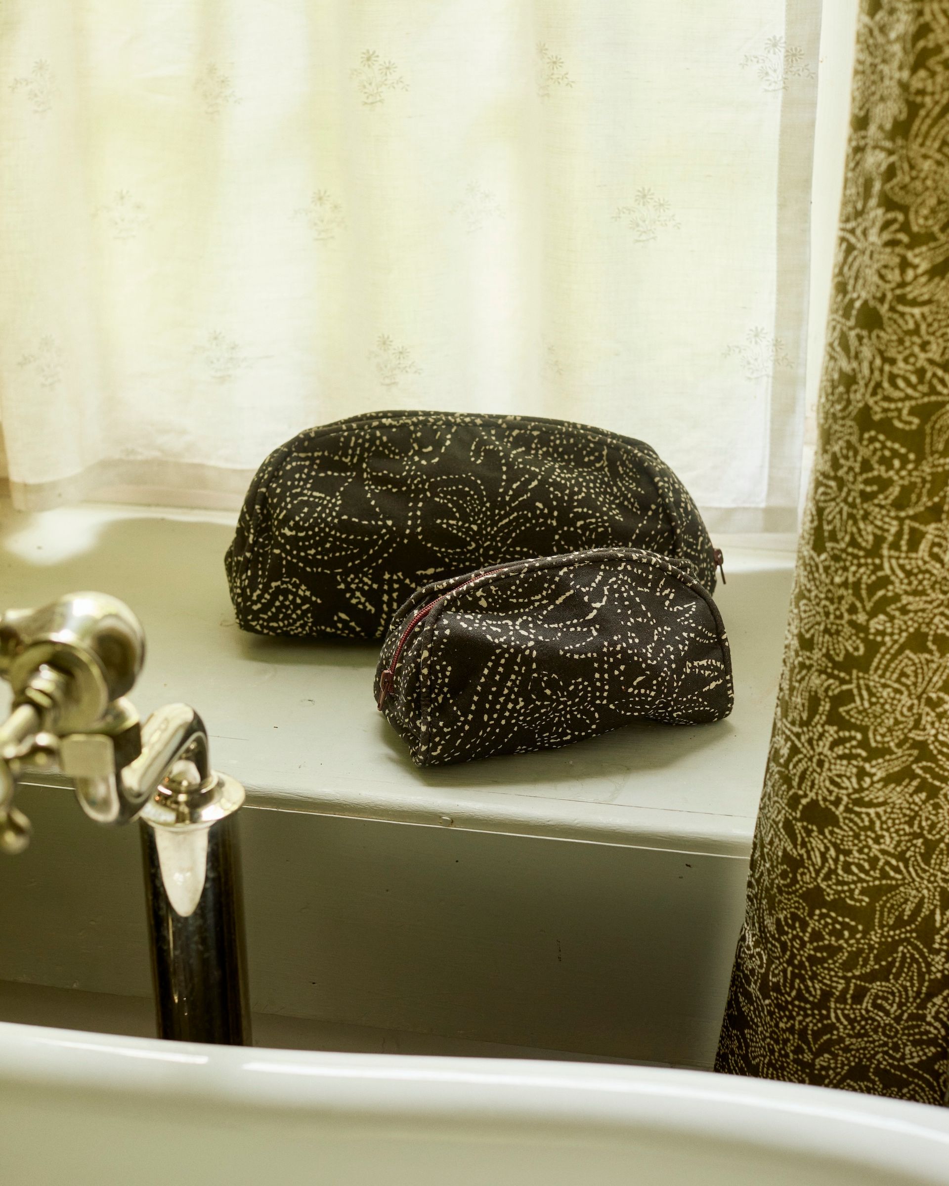 two toilers bags sit on a green window sill next to the bath tap and printed curtain