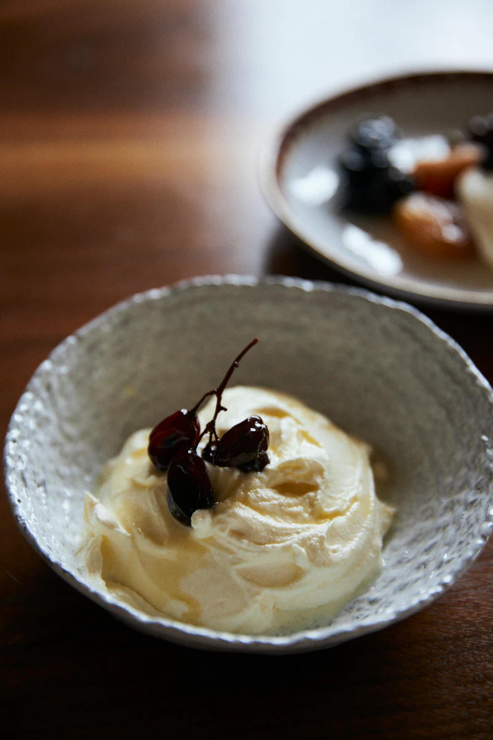 Textured bowl with creamy dessert topped with dark cherries. Blurred dish in background on wooden surface.