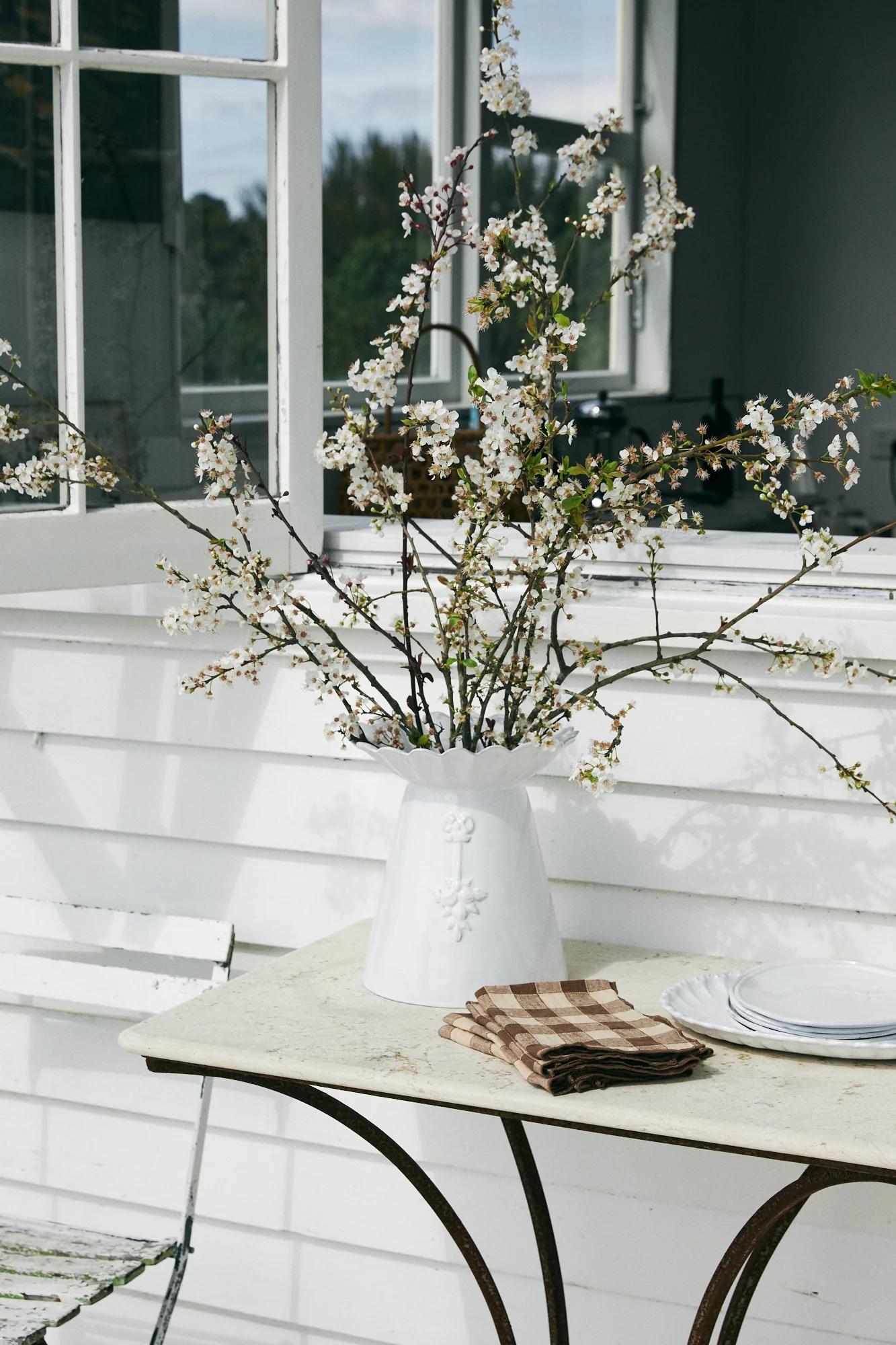 White vase with blossoms on rustic table. Checkered napkins and Astier de Villatte plate nearby. Open window shows green background.