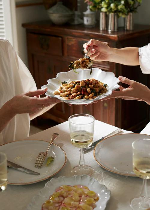 Two people share a meal at a table set with wine glasses, plates, utensils and sliced radishes. One person serves chickpeas and greens whilst the other holds a dish.