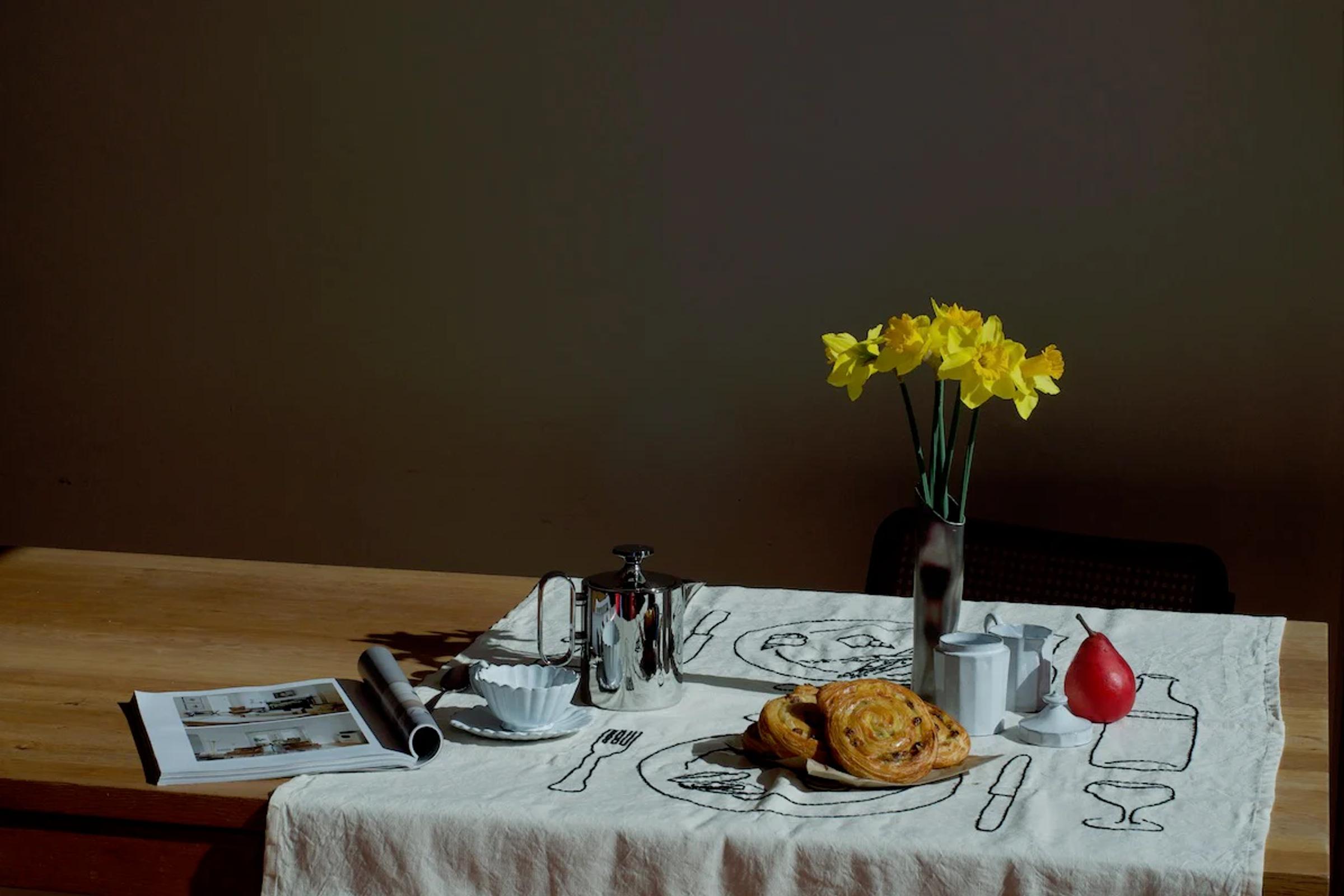 Wooden table with embroidered cloth, coffee pot, cup, saucer, pastries, yellow flowers. Red pear and open magazine.