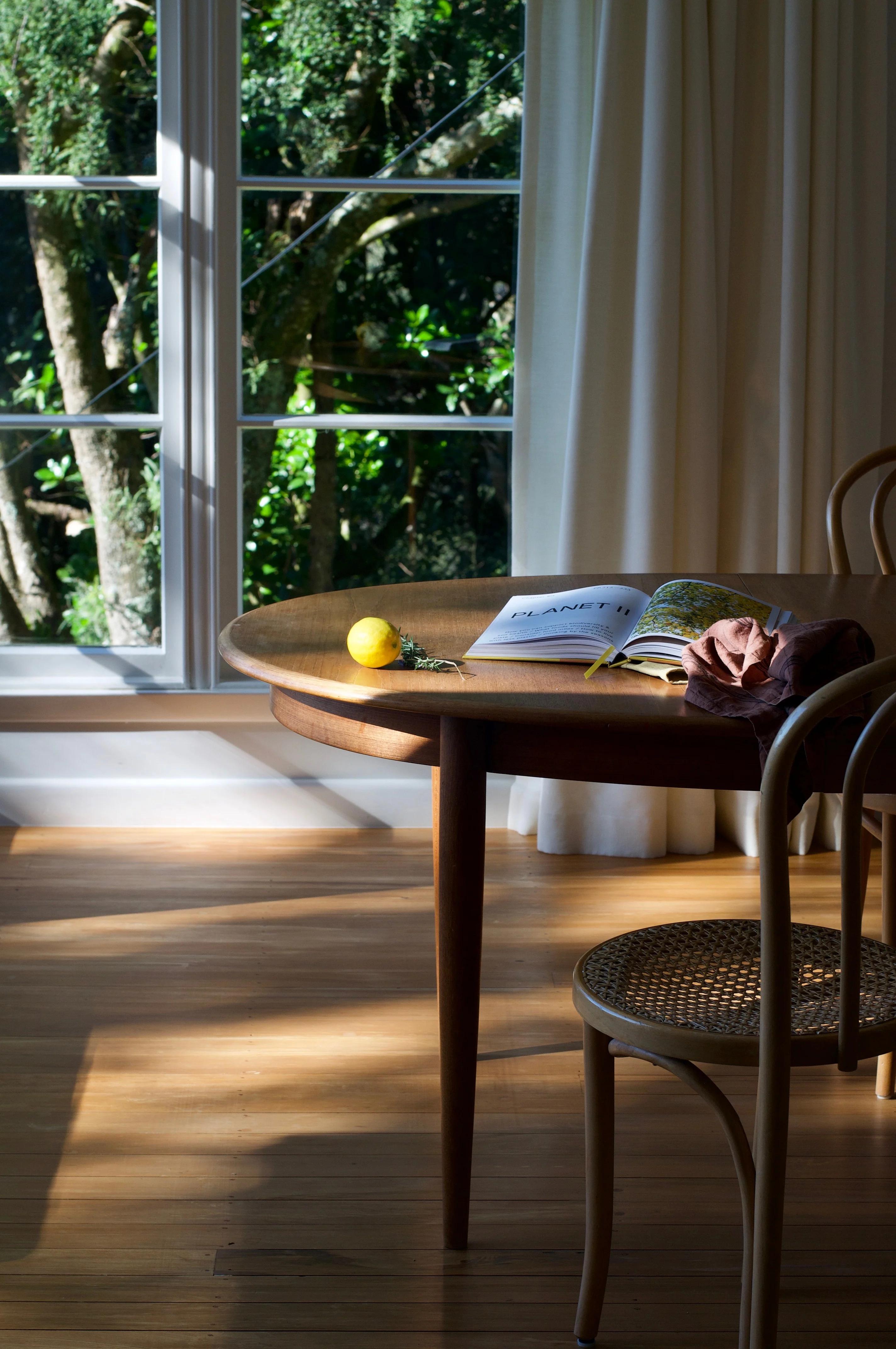 Wooden table with open book, lemon, fabric in sunlit room. Wooden chair nearby. Shadows on floor, greenery outside.