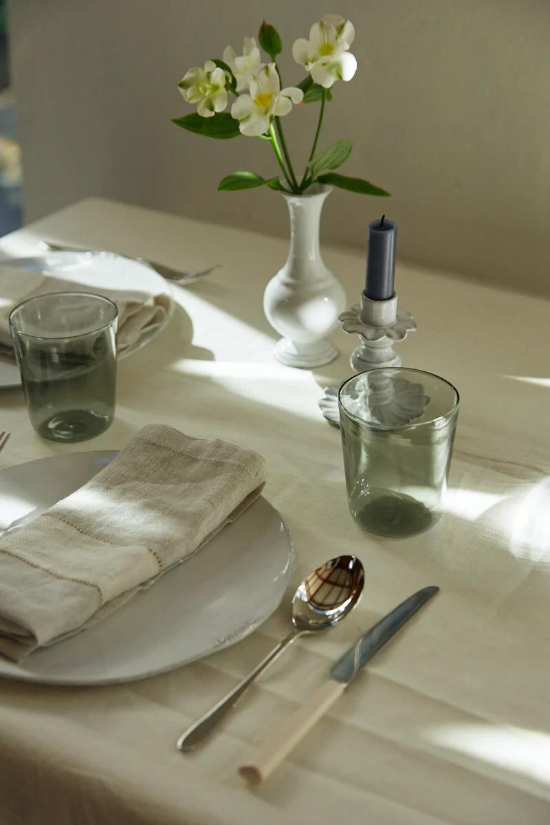 Table set with white cloth, green glasses, beige Libeco napkins. White flowers and blue candle. Minimalist, elegant setup in soft sunlight.