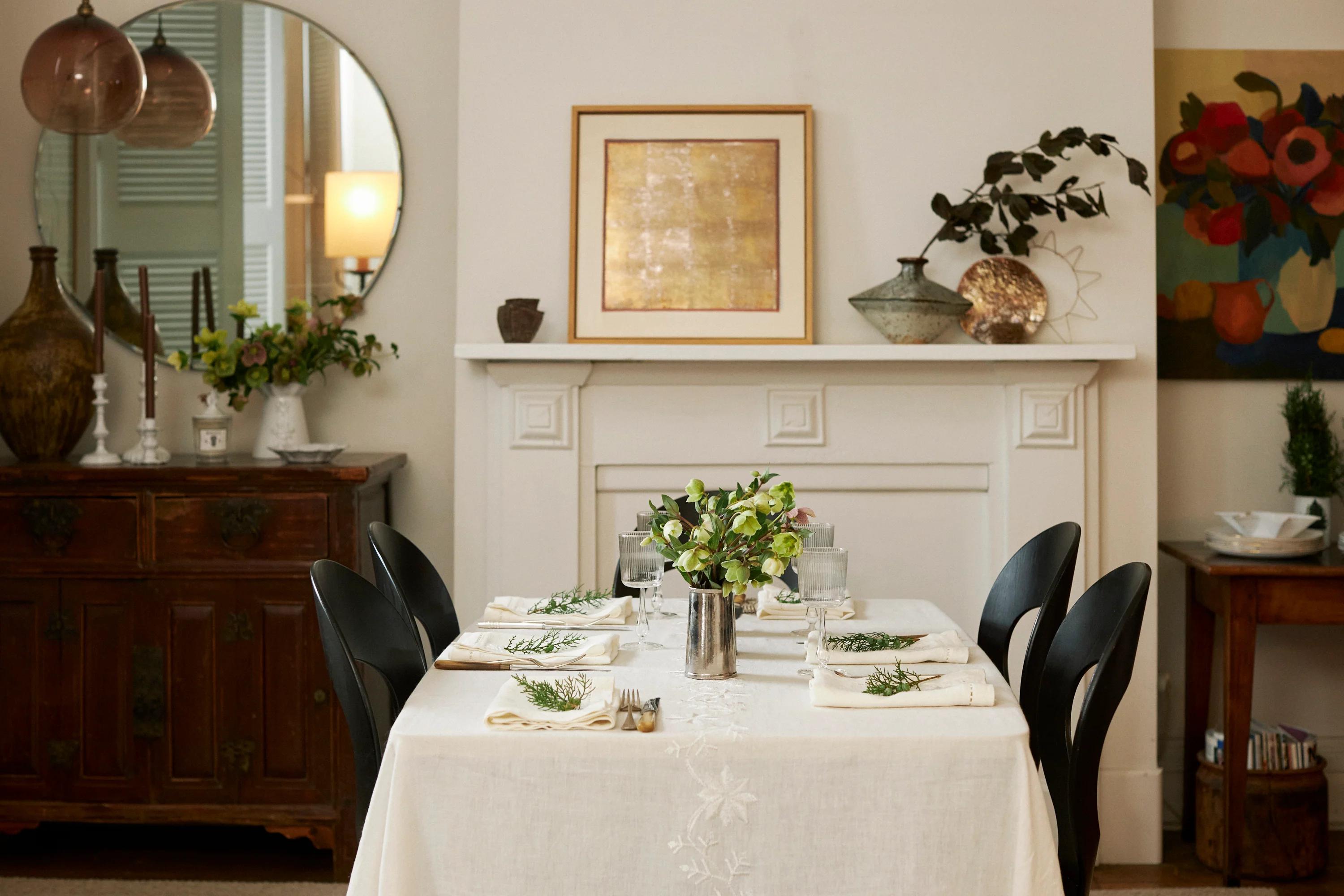 A cosy dining room with a set table, decorated fireplace, and vintage sideboard. Artwork and floral centrepieces create a festive, elegant atmosphere.