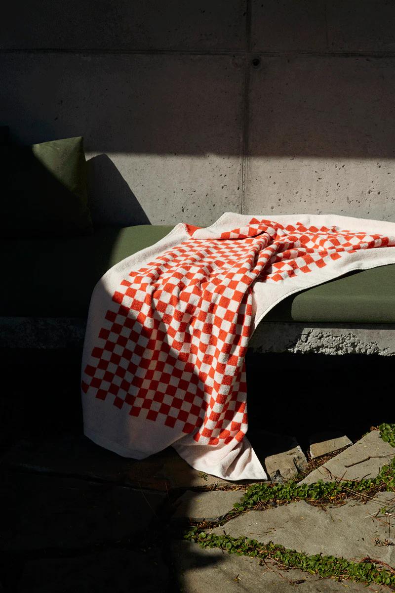 A red and white checkered blanket draped over a gray bench seat with a green cushion, partially in sunlight. A concrete wall and stone pavers with greenery are visible in the background.