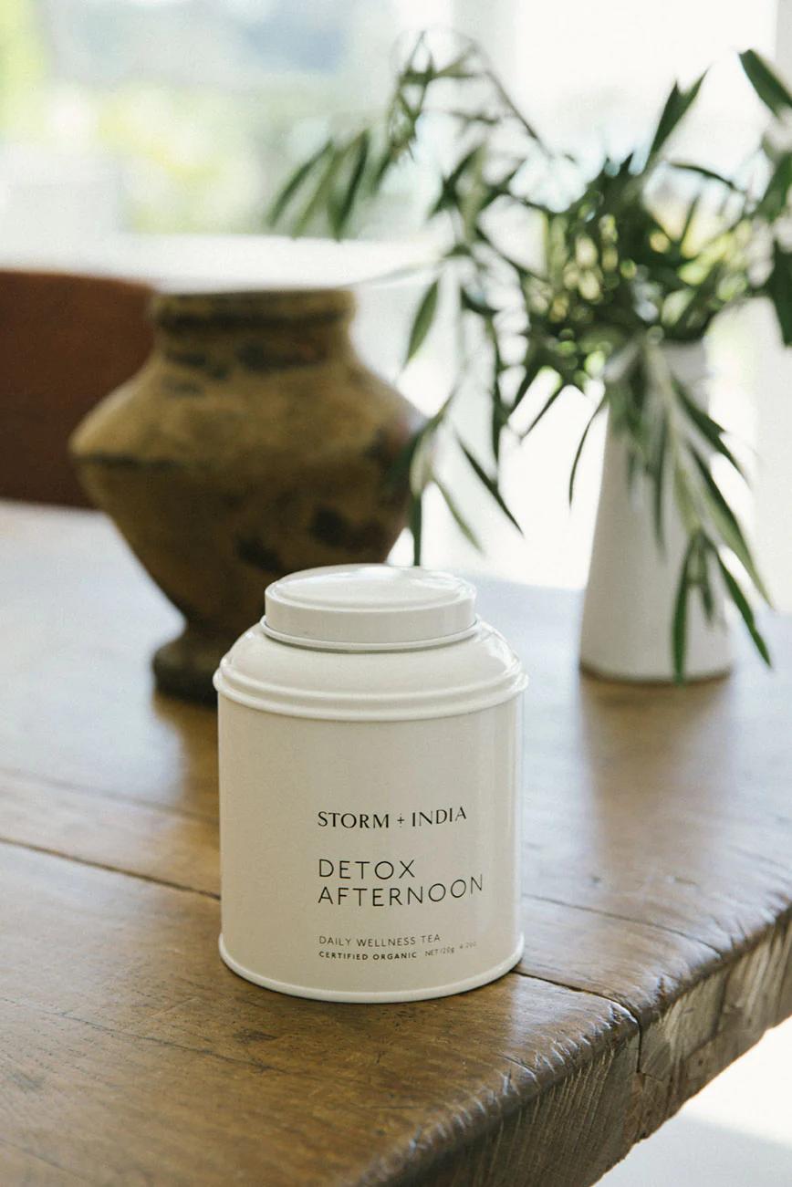 White "STORM + INDIA DETOX AFTERNOON" tea canister on wooden table. Clay and white vases with foliage in background near sunlit window.