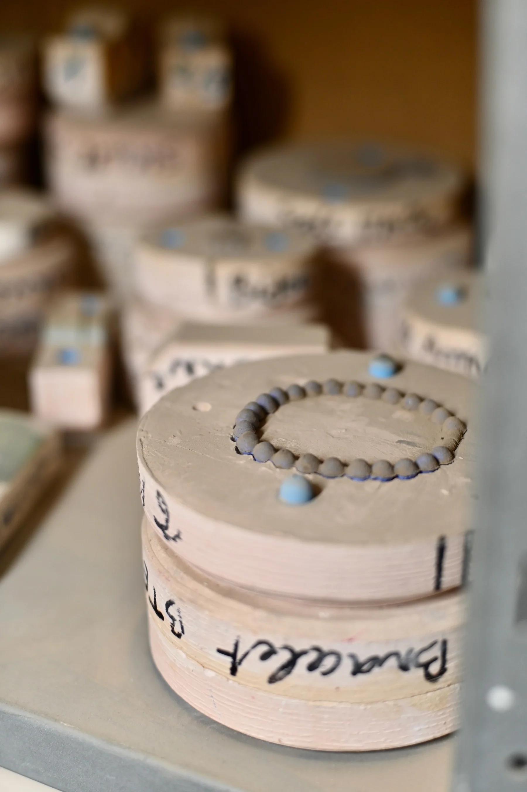 Ceramic or plaster molds of various sizes on shelves. Prominent circular mold with small round beads in foreground. Some molds labeled with handwritten numbers.