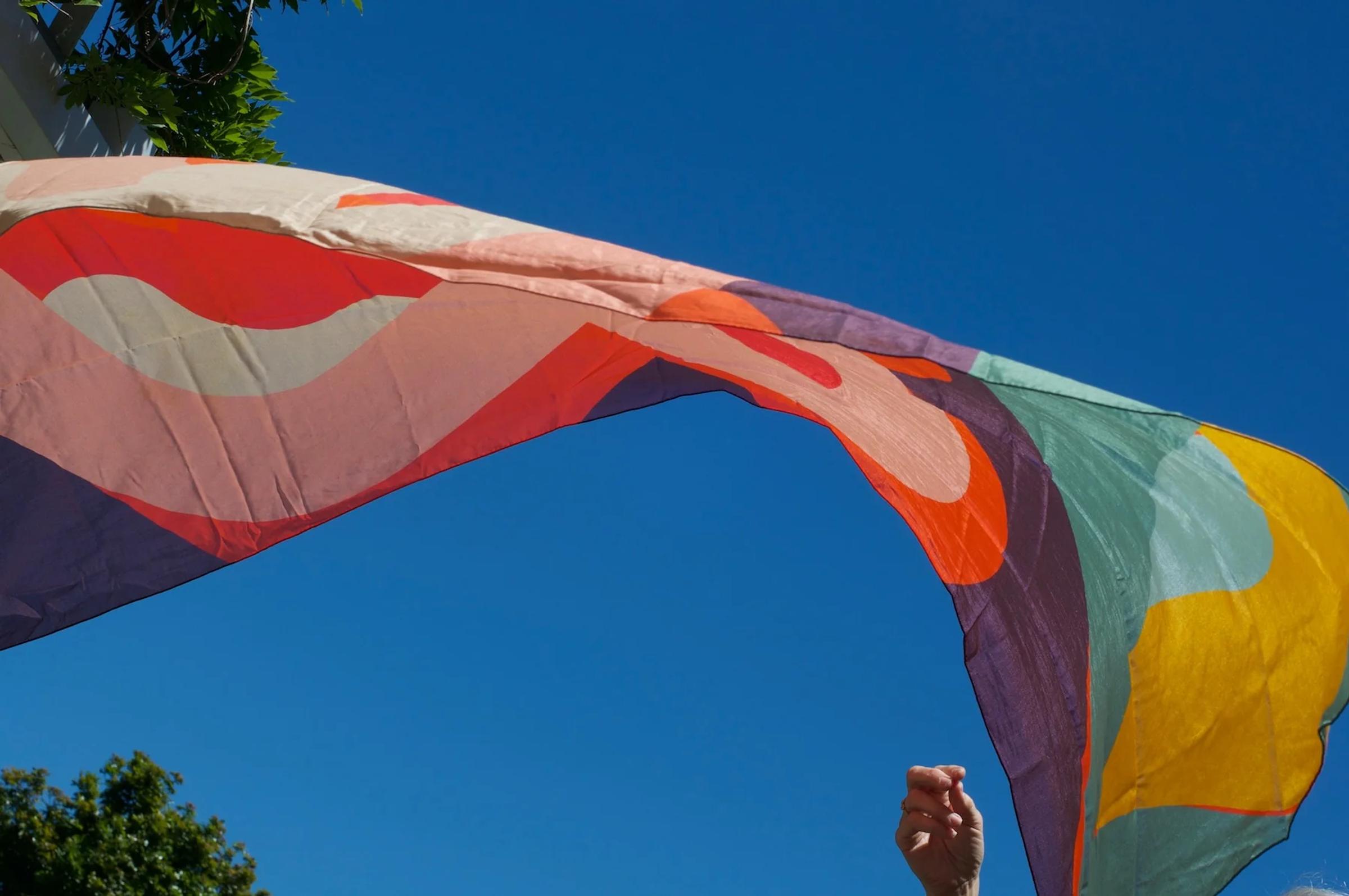 Colourful kite flying in blue sky. Tree and house roof visible. Person holding kite string, others watching below.