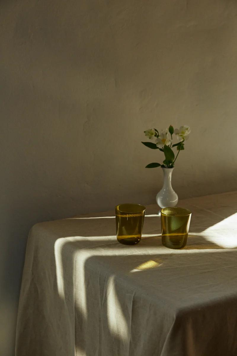 Belgian linen-covered table with green glass cups and small white vase of flowers. Warm sunlight casts shadows.