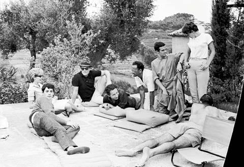 Black and white image of eight people on outdoor patio. Some lounging, others standing. Trees in background.