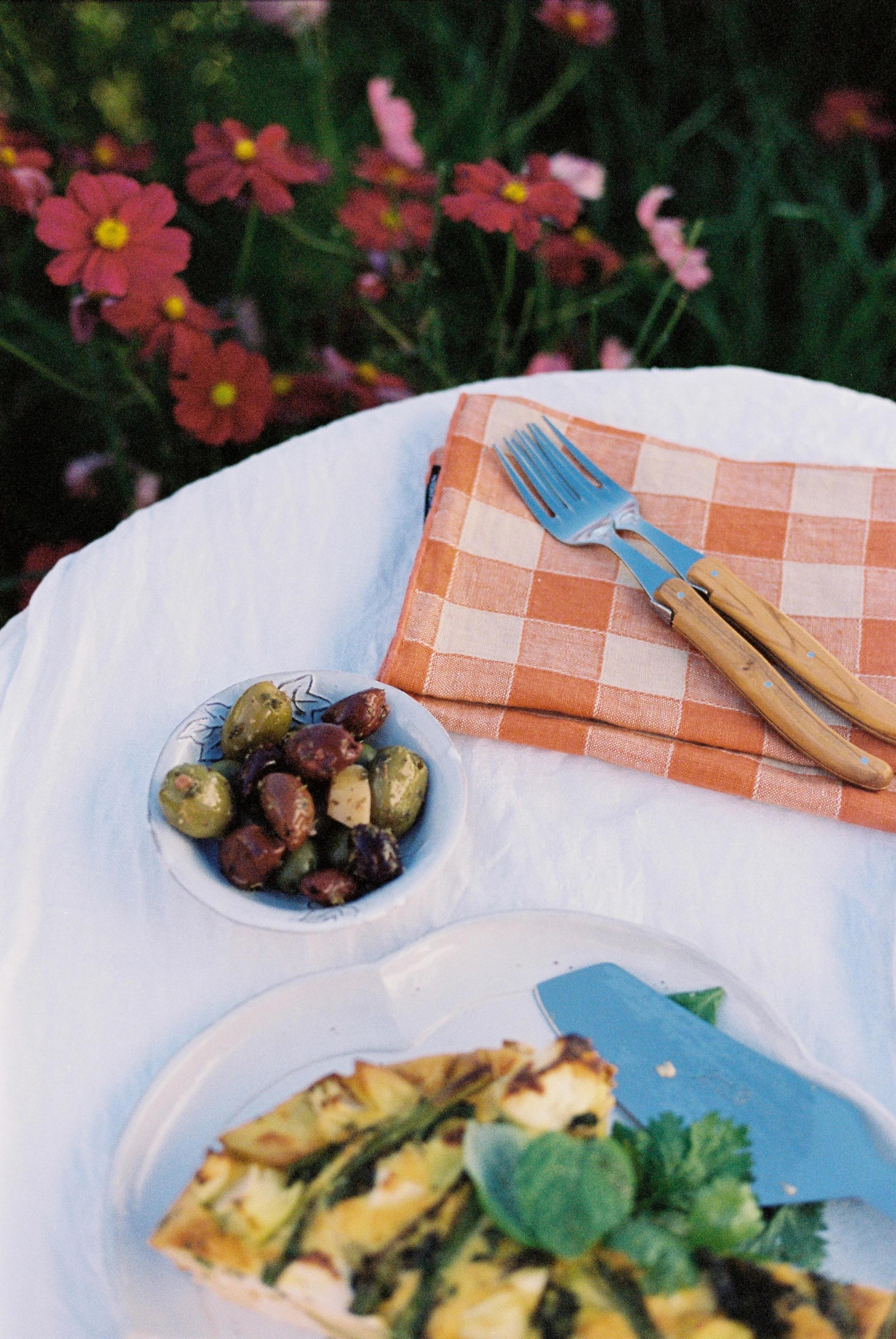 Table with orange napkin, wooden cutlery, green frittata slice, and mixed olives. Red flowers and green foliage in background create sunny garden feel.