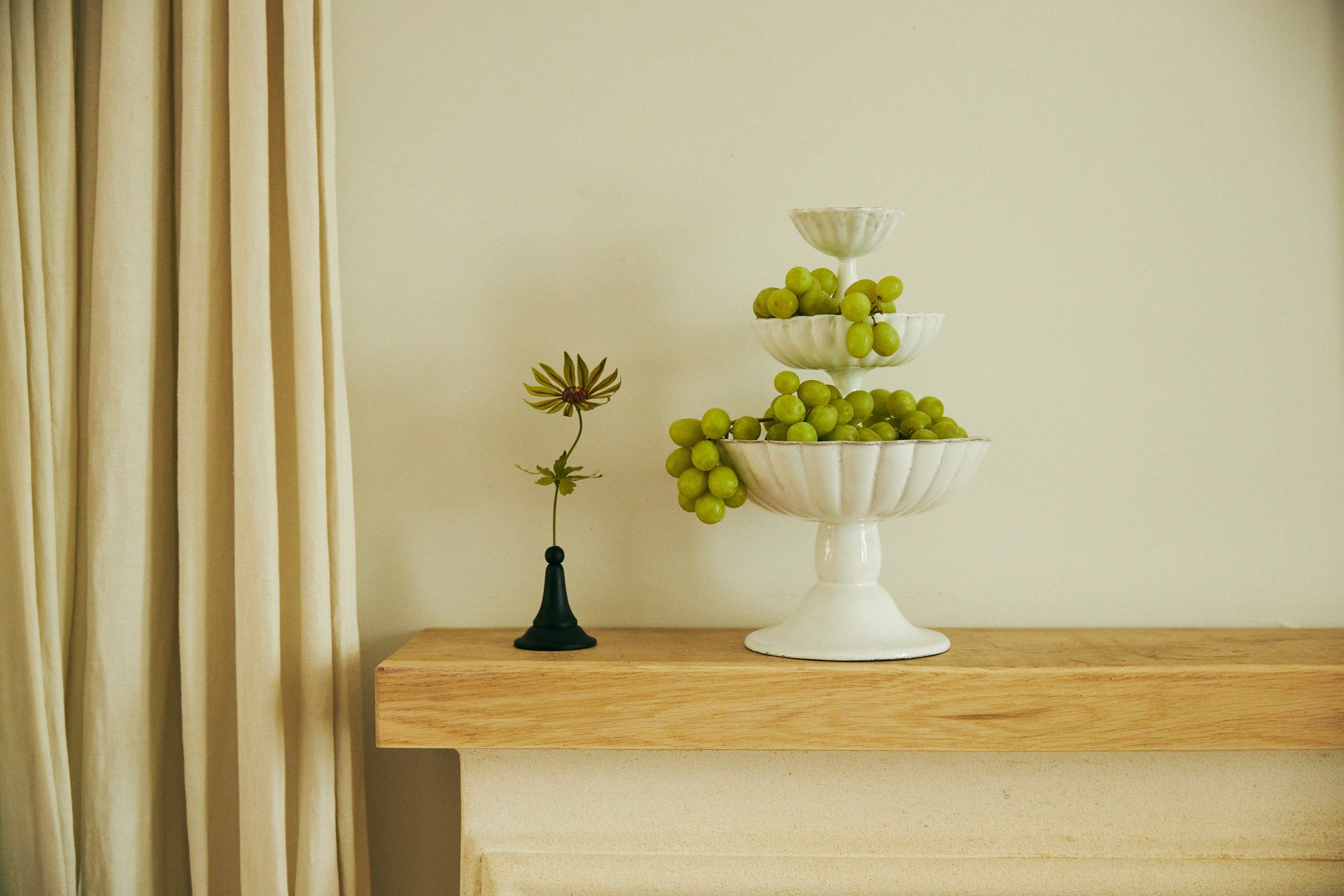 White tiered dish with grapes on wooden mantel. Black vase with single flower. Light curtain and beige wall background.