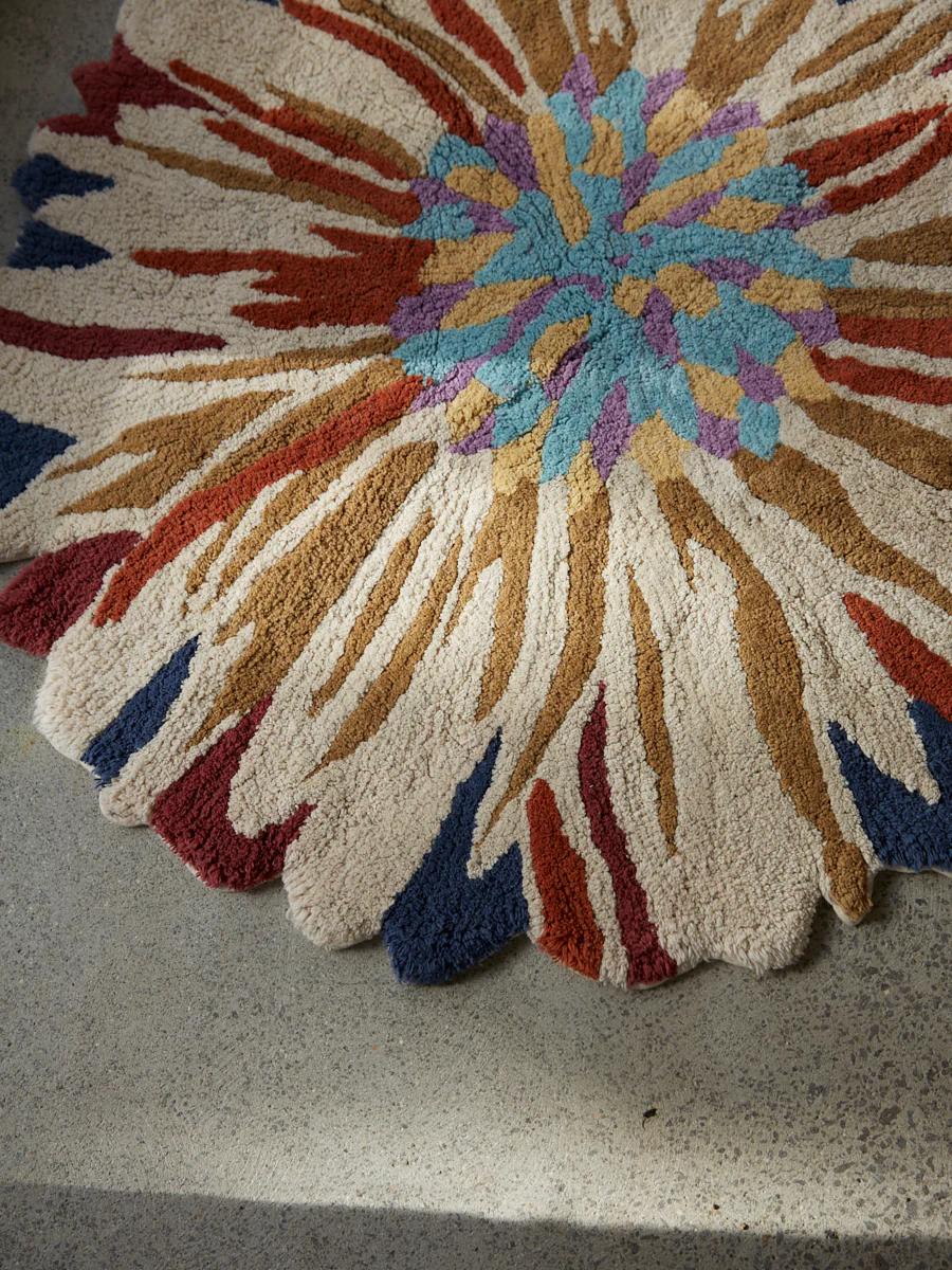 A round, textured rug with a multicoloured flower design in earthy tones. Made from soft velour, it sits on concrete with sunlight casting a faint shadow.