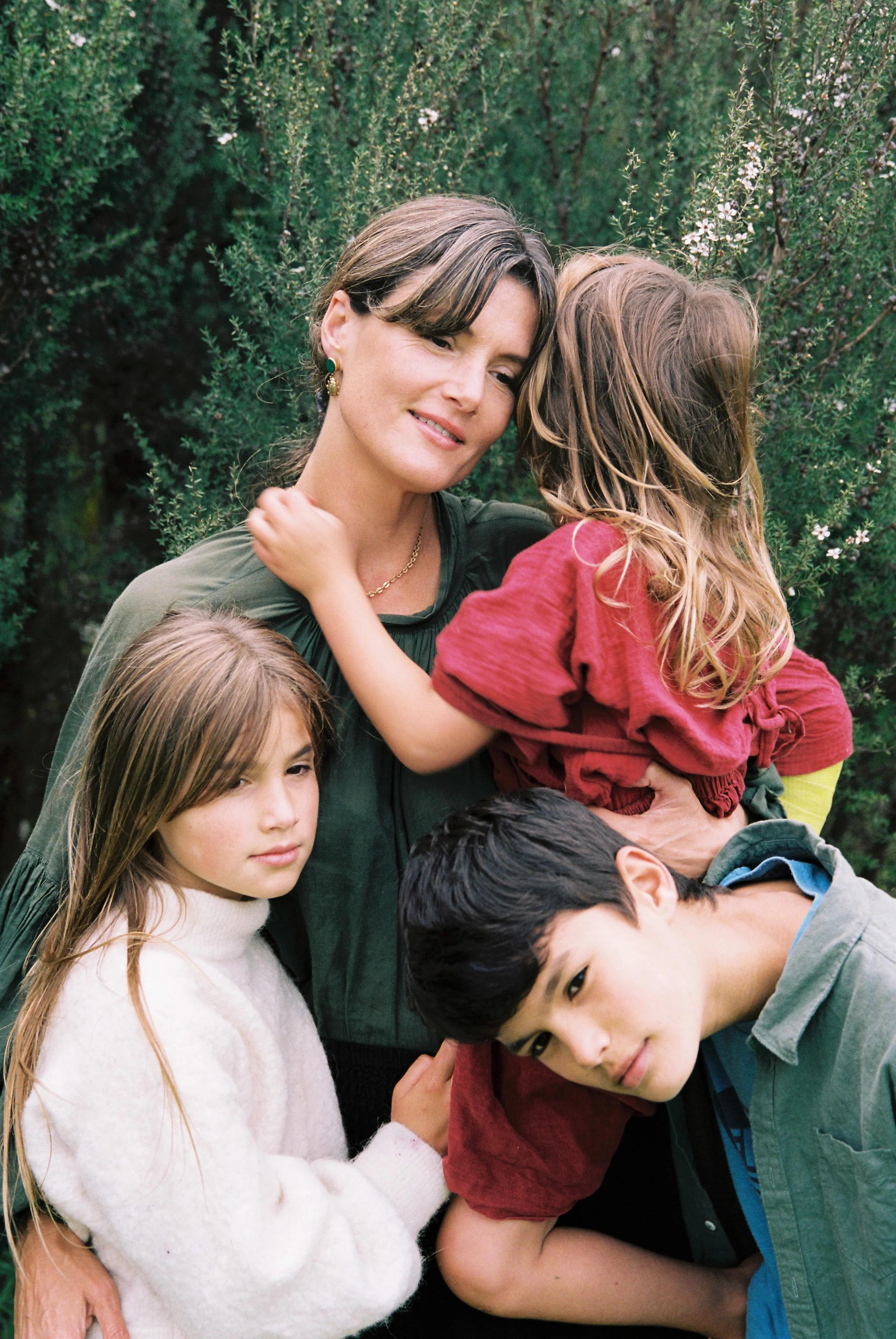 A woman with three children embraces them under a tree. One child in a cream sweater stands beside her, another in green leans on her, and a third in red rests on her shoulder.