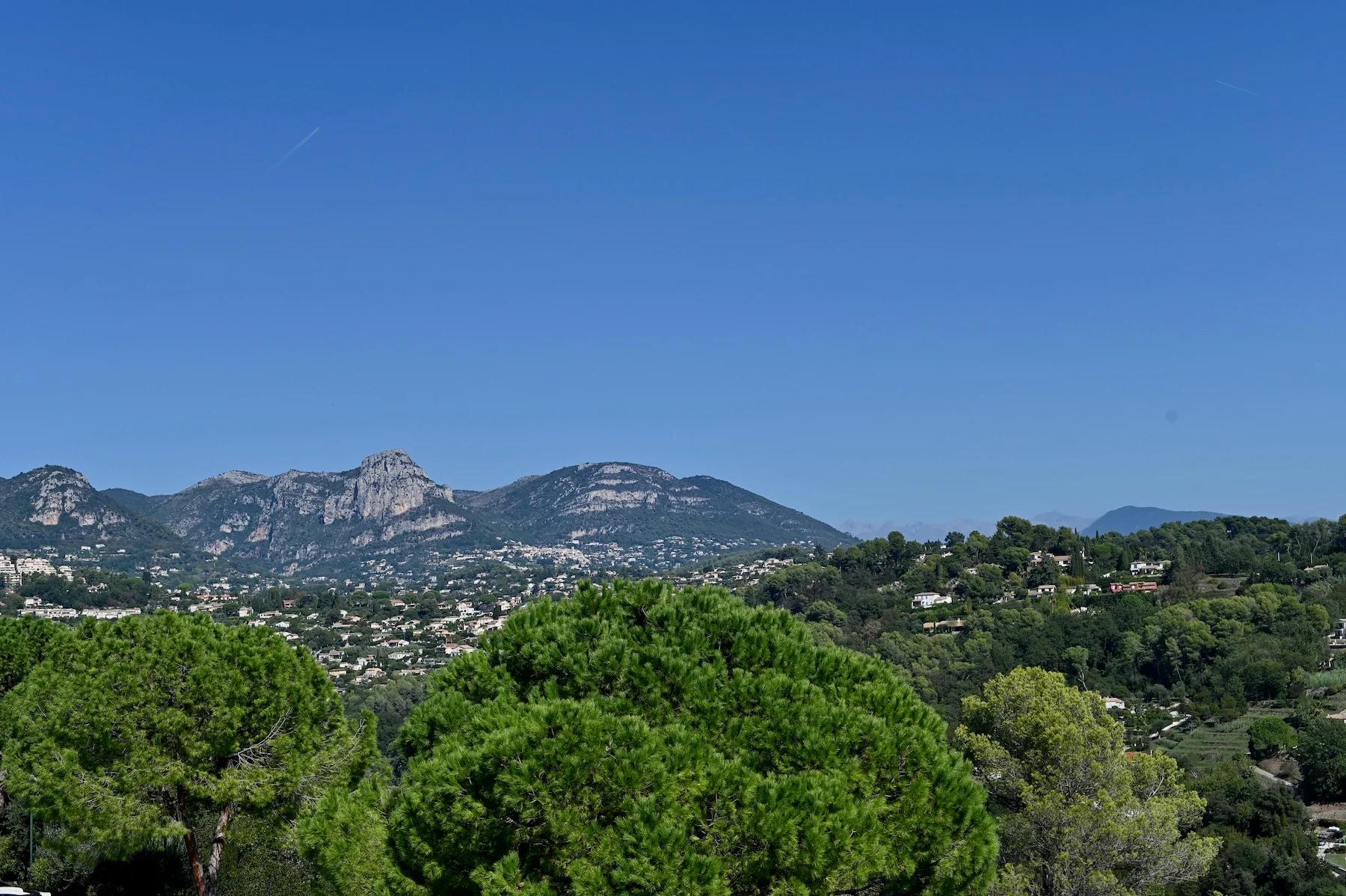 Panoramic view of lush, hilly South of France landscape. Green trees in foreground, mountains under blue sky, with scattered houses nestled among trees.