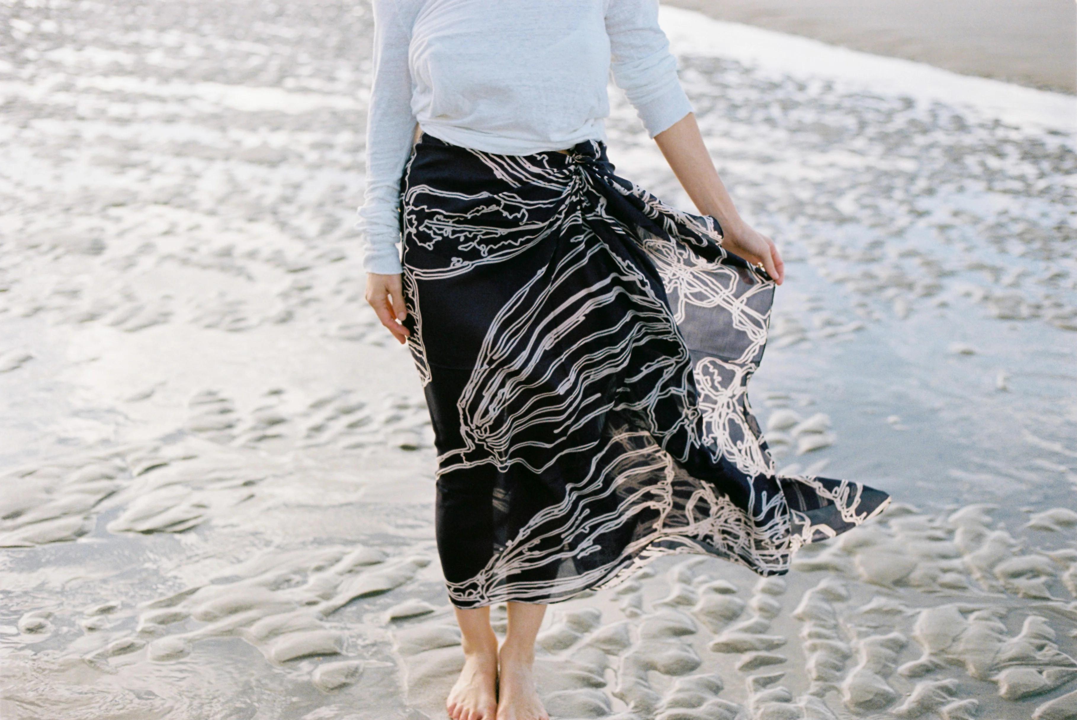 Person in long black and white patterned skirt and light top on wet, sandy beach. Wind catching skirt. Face not visible.