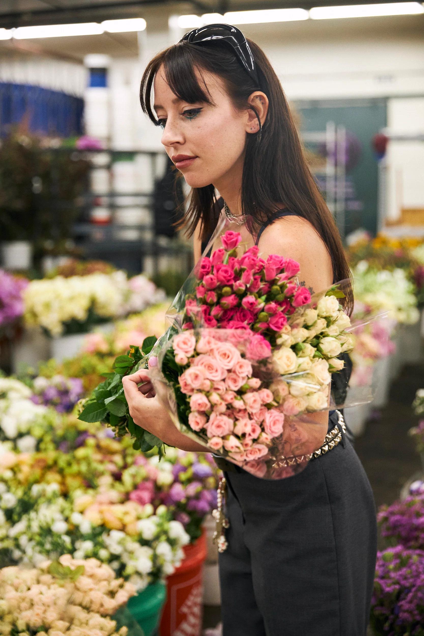 Woman with long brown hair holds pink and white roses in flower shop. Colorful arrangements and plants surround her.