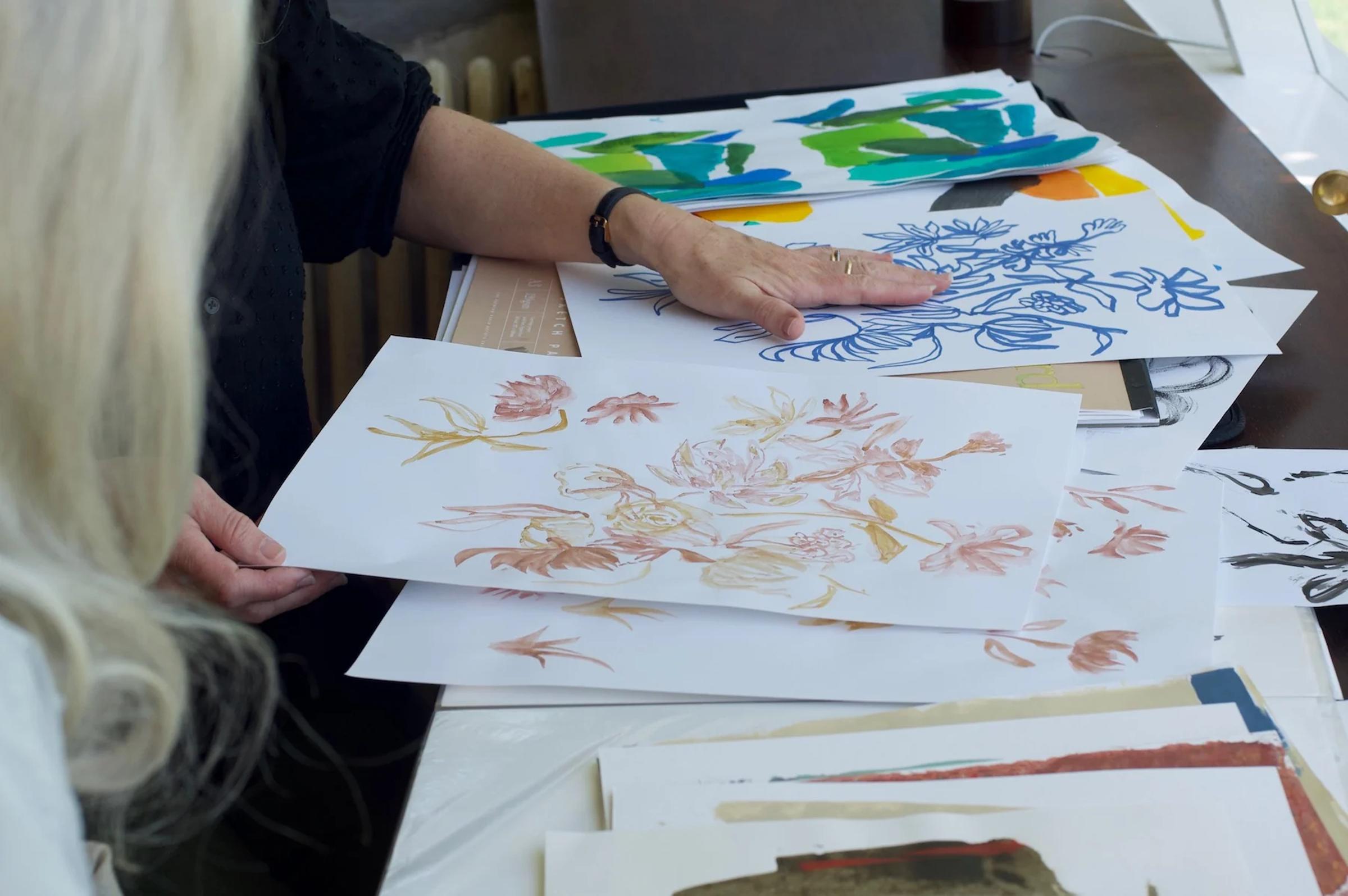 Two people reviewing colourful floral and abstract prints on table. Hand holding pink flower print visible.
