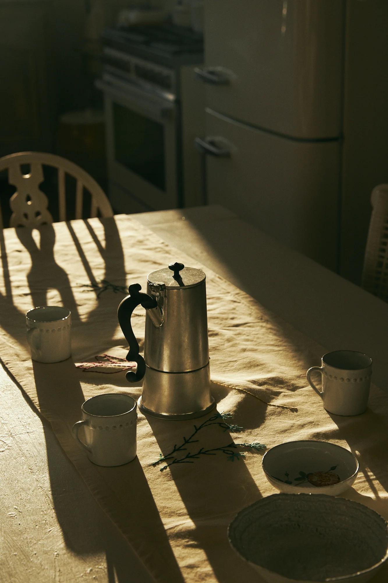 Sunlit kitchen table with espresso pot, cups, and bowl. Floral tablecloth, vintage appliances. Shadows create serene holiday breakfast scene.