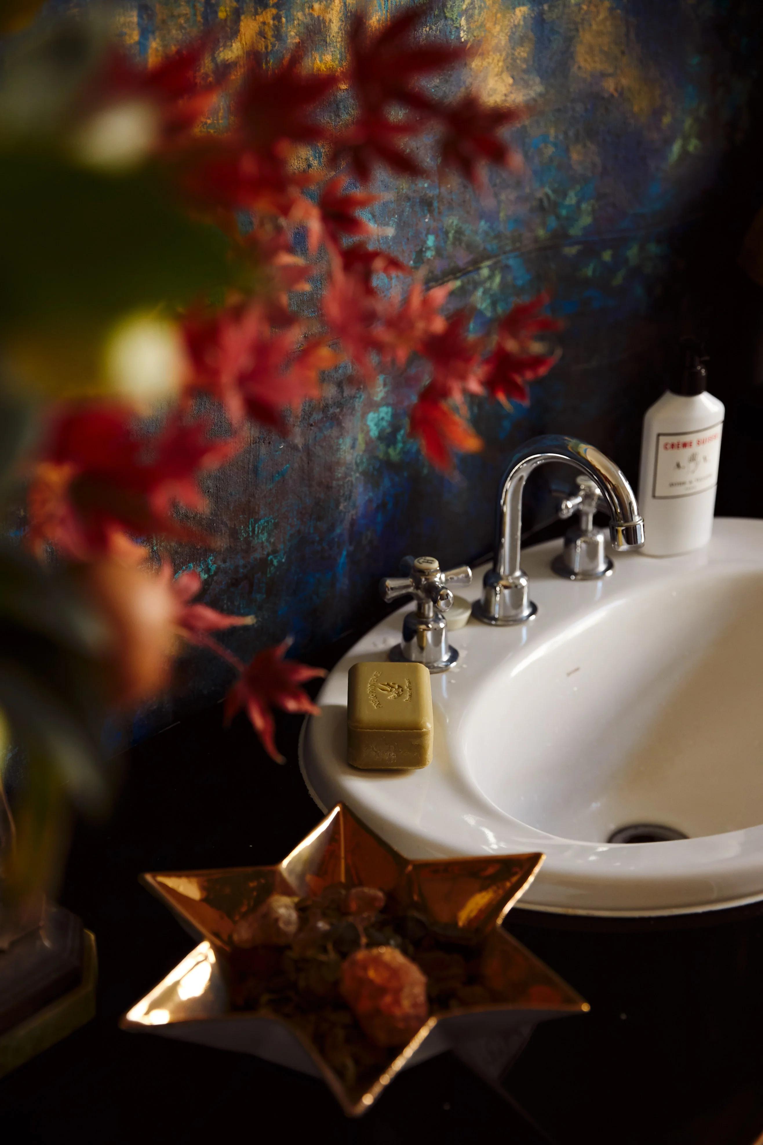 Elegant bathroom with silver faucet, lotion, and soap. Star-shaped dish holds decorative items. Red flowers and blue-gold wall evoke winter solstice warmth.