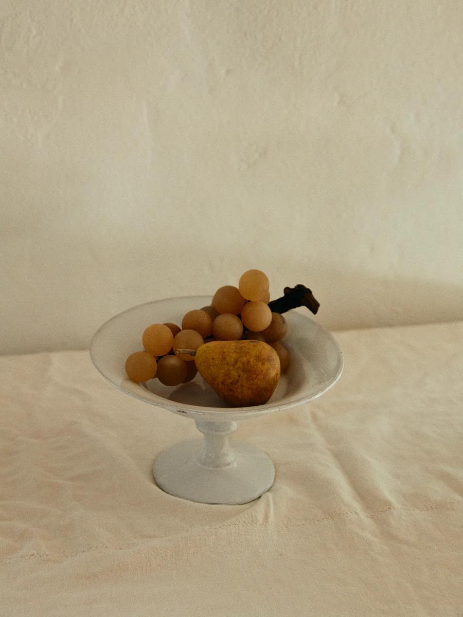 White ceramic bowl on pedestal holds yellow grapes and pear. Light, neutral background creates simple, rustic aesthetic.