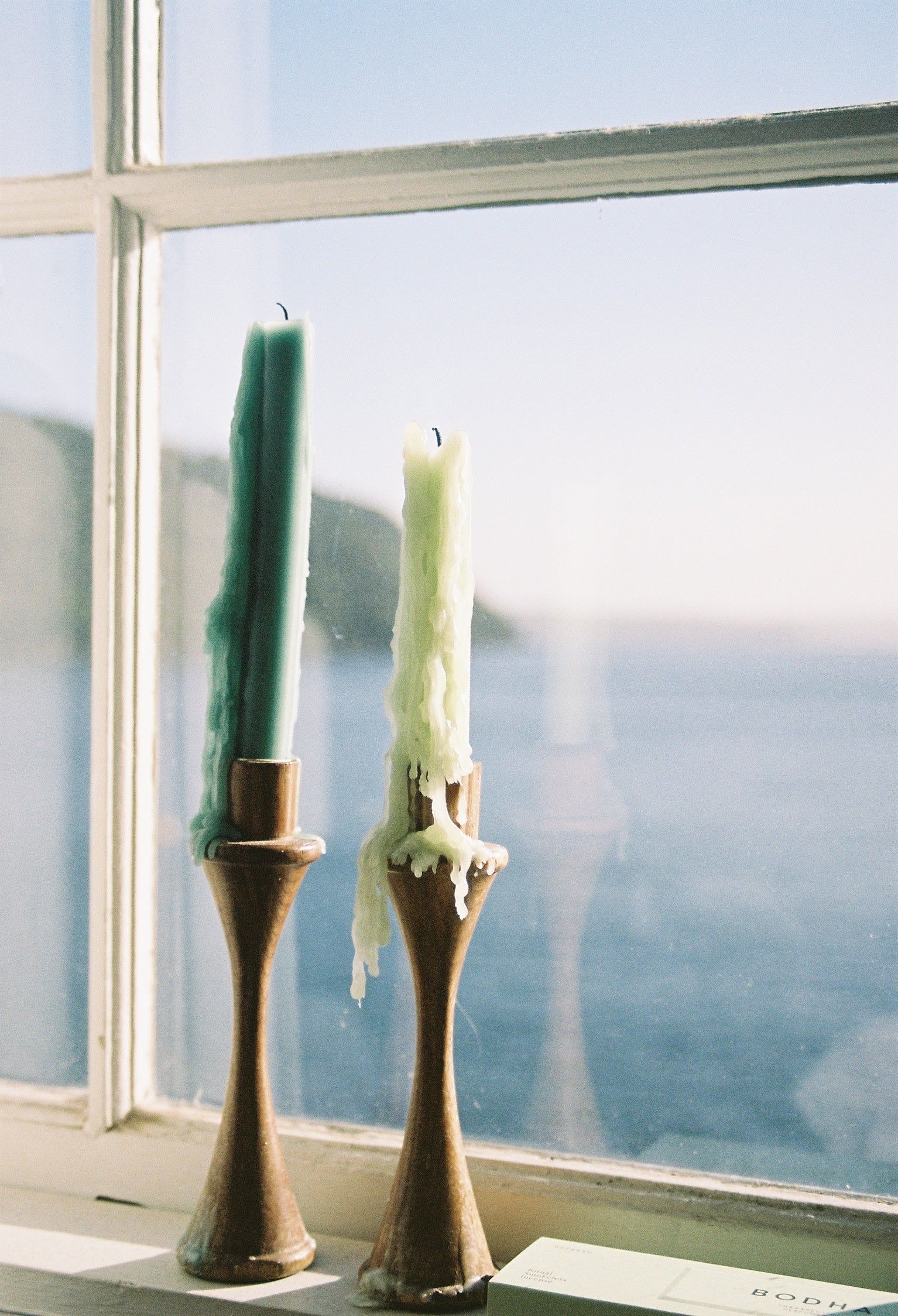 Two tall candles, one green and one white, placed in wooden holders on a windowsill, with a seaside view of waves and a distant boat shed.