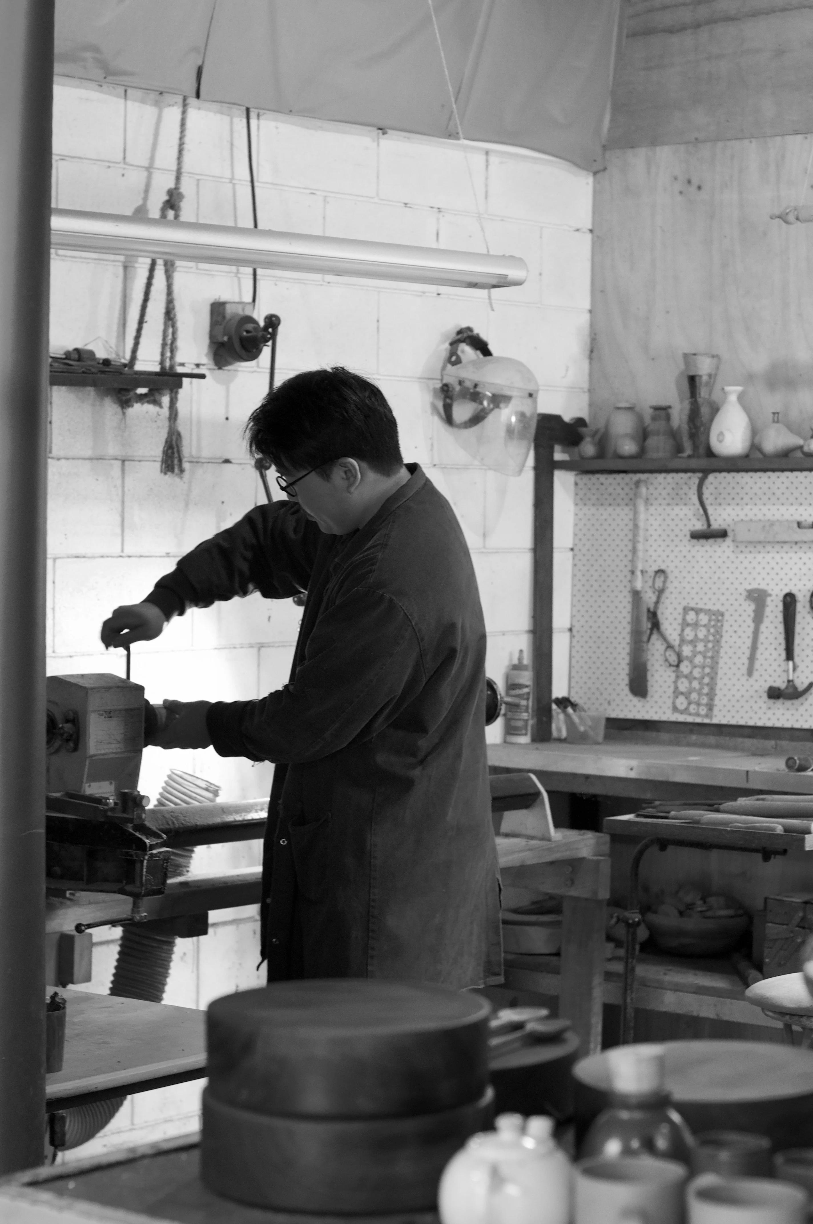 Person in long coat works on equipment in industrial workshop. Shelves and benches filled with tools and materials reflect NZ wood craftsmanship.