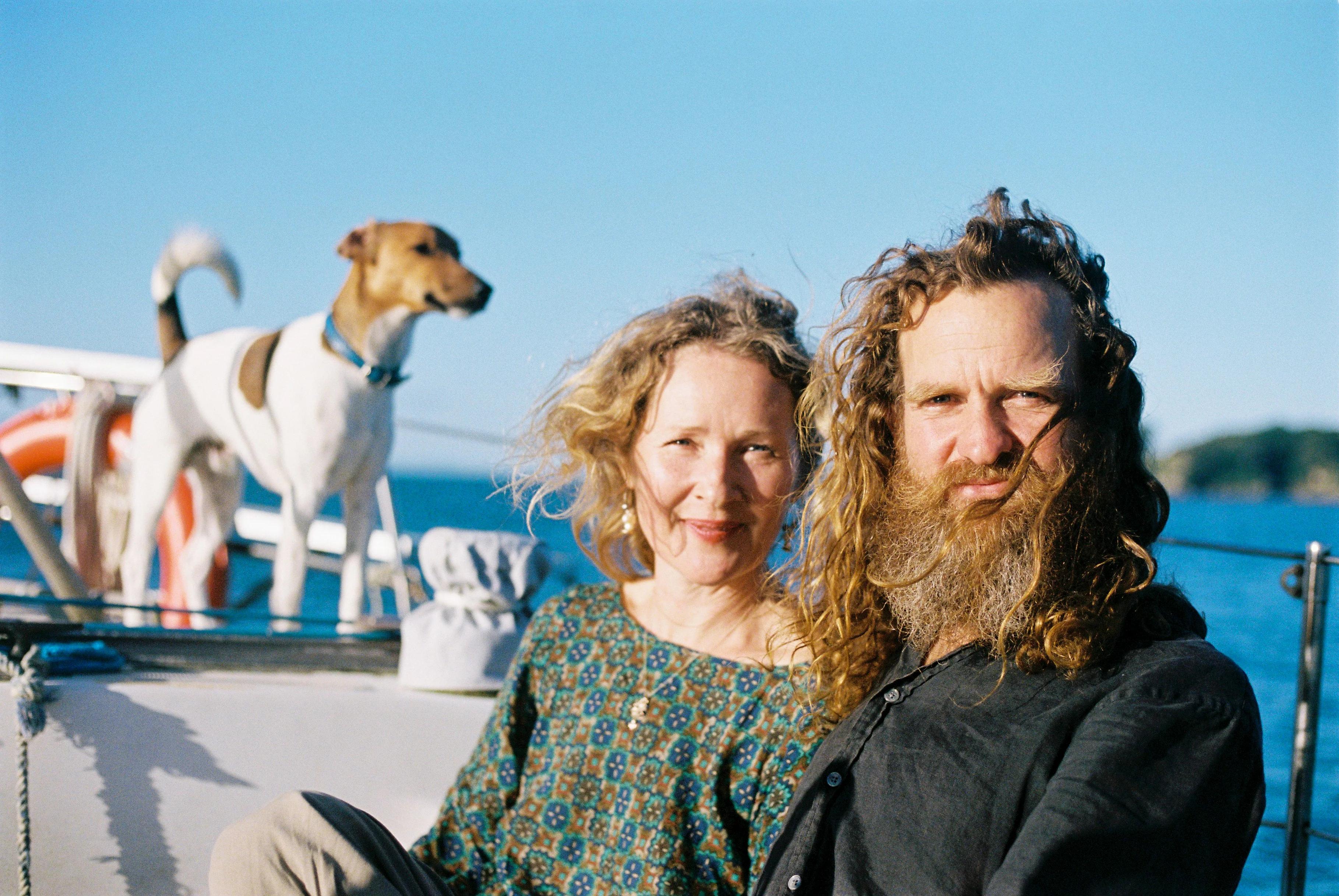 Blonde woman, bearded man, and dog on boat under blue skies. Waiheke Island waters visible.