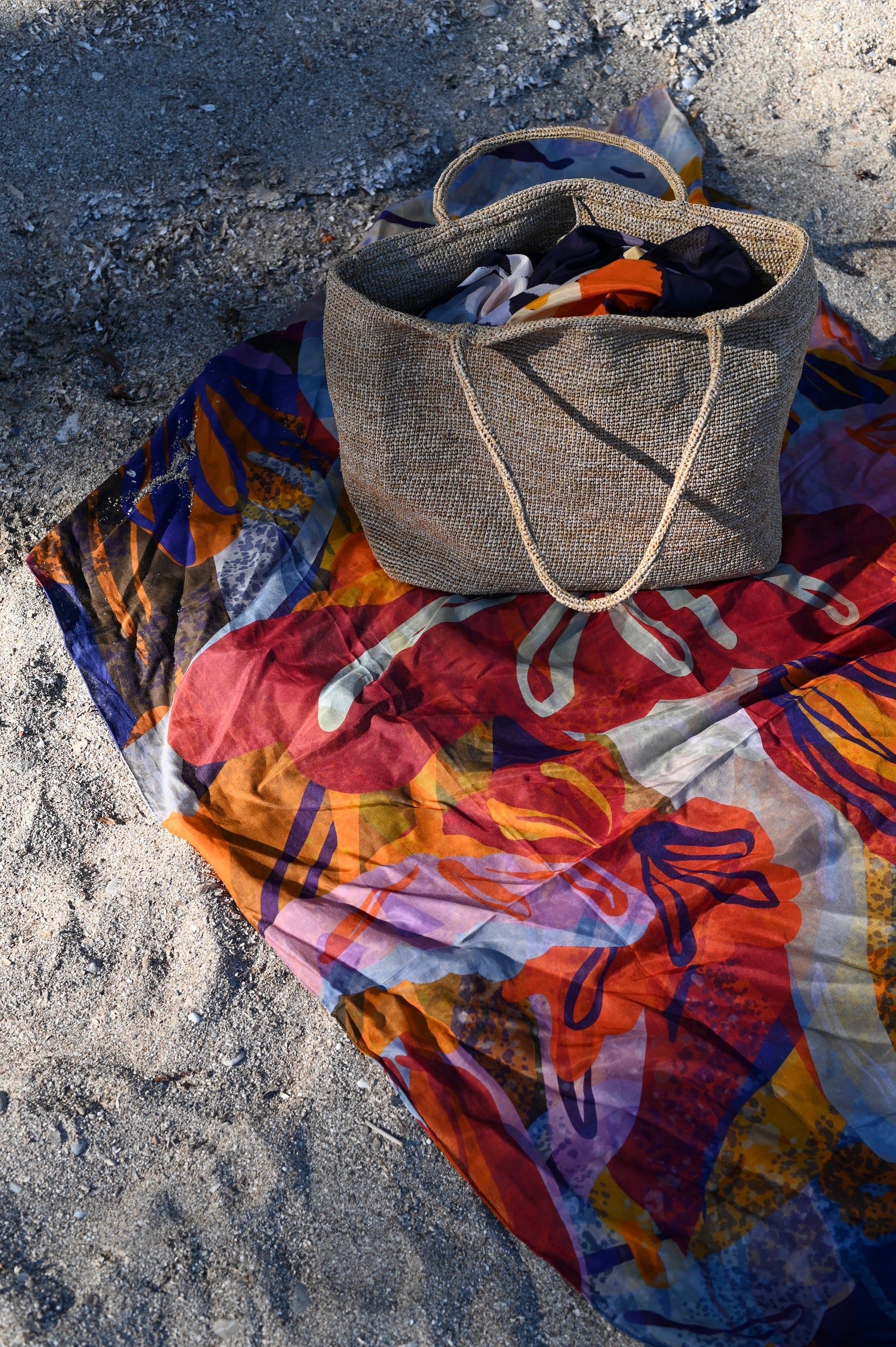 Colourful patterned beach towel on sand. Beige textured beach bag filled with items, including sarongs, on top.