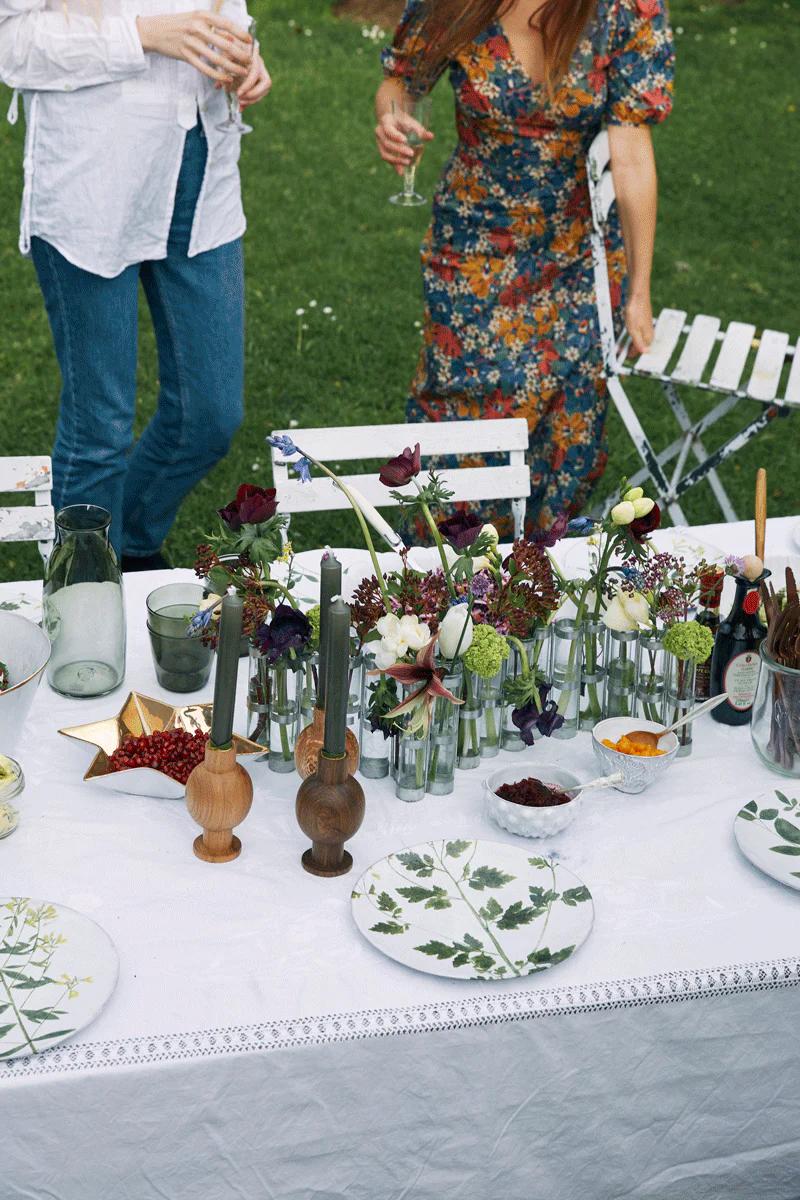 White-clothed table with patterned plates and flowers. Two people nearby, one in white top, other in floral dress.