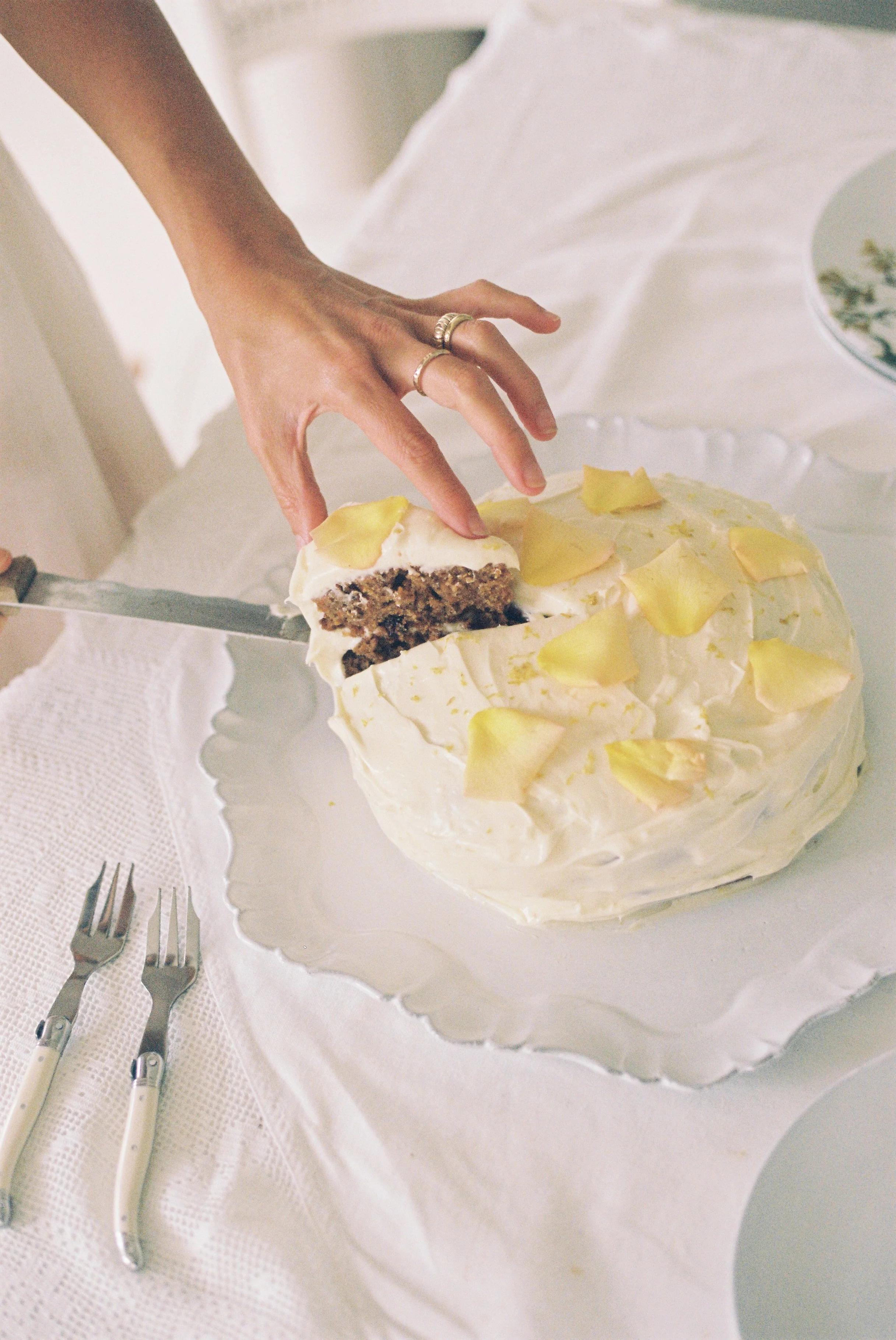 Person slicing frosted carrot cake on white plate. Light icing, yellow garnish. Forks on white cloth nearby.