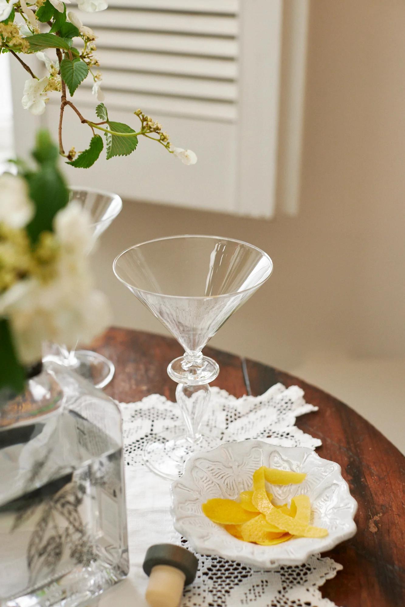 Two empty martini glasses, a vodka bottle, and a bowl of lemon twists on a wooden table. A lace doily and flowers in the background create a sophisticated atmosphere.