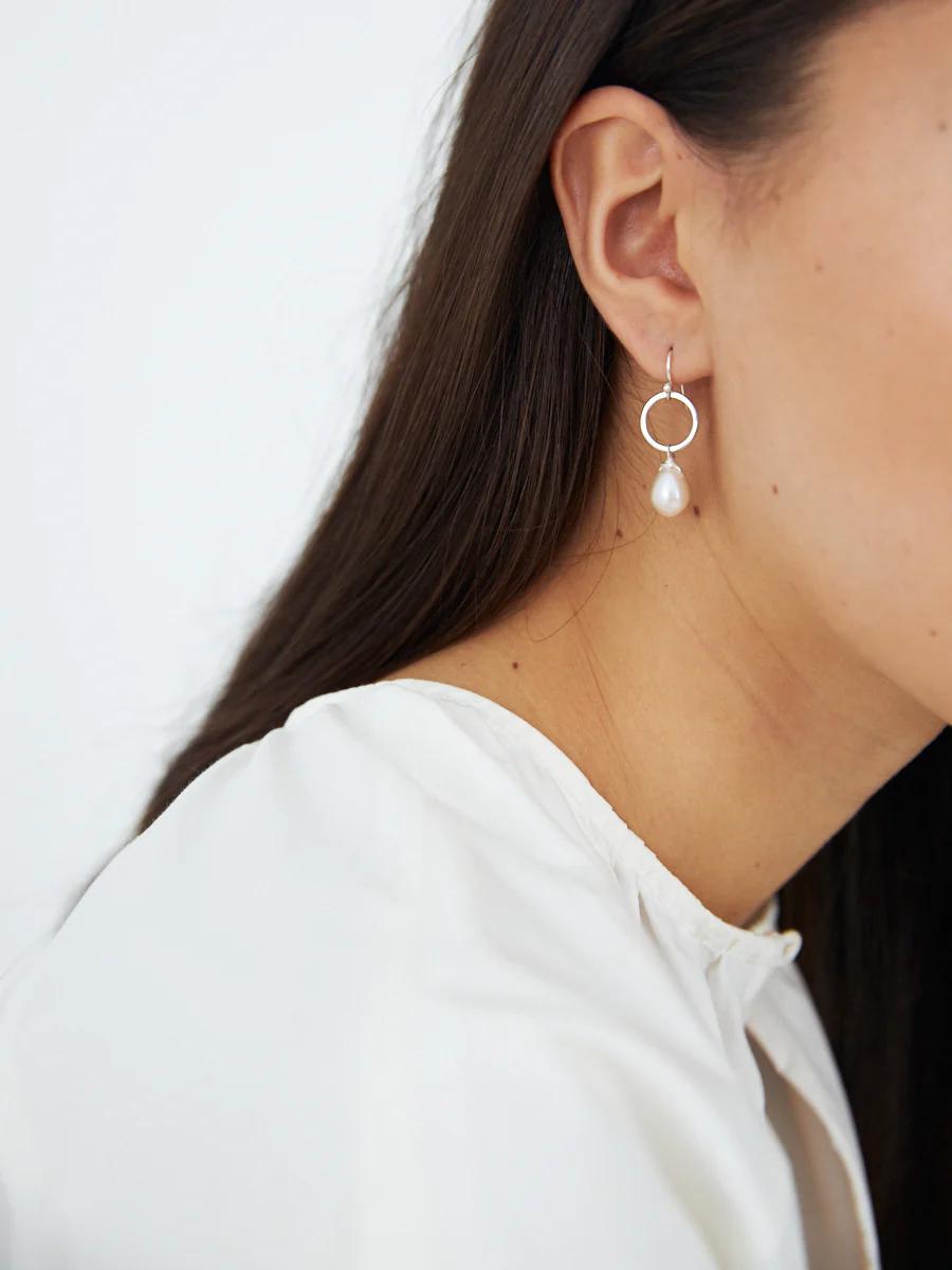 Close-up of person with long, dark hair wearing white top and silver hoop earring with hanging pearl.