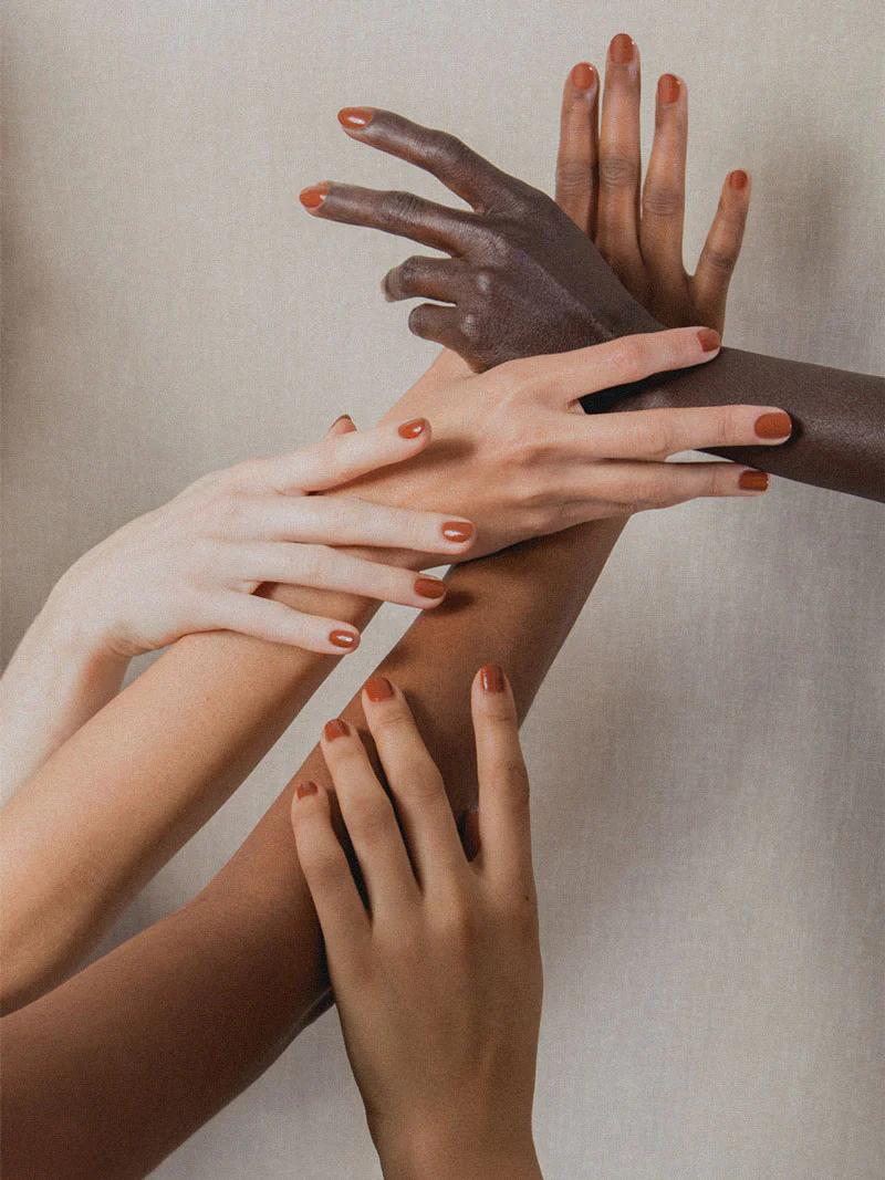 Four hands with painted nails in reddish-orange intertwine on a neutral background, showcasing unity and diversity. The polish reflects J. Hannah’s commitment to elegance and inclusivity.