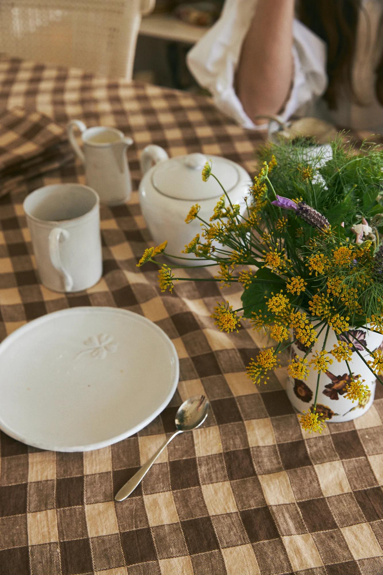 Checkered tablecloth with white plate, teaspoon, mug, teapot, milk jug. Flower vase nearby. Person partially visible.